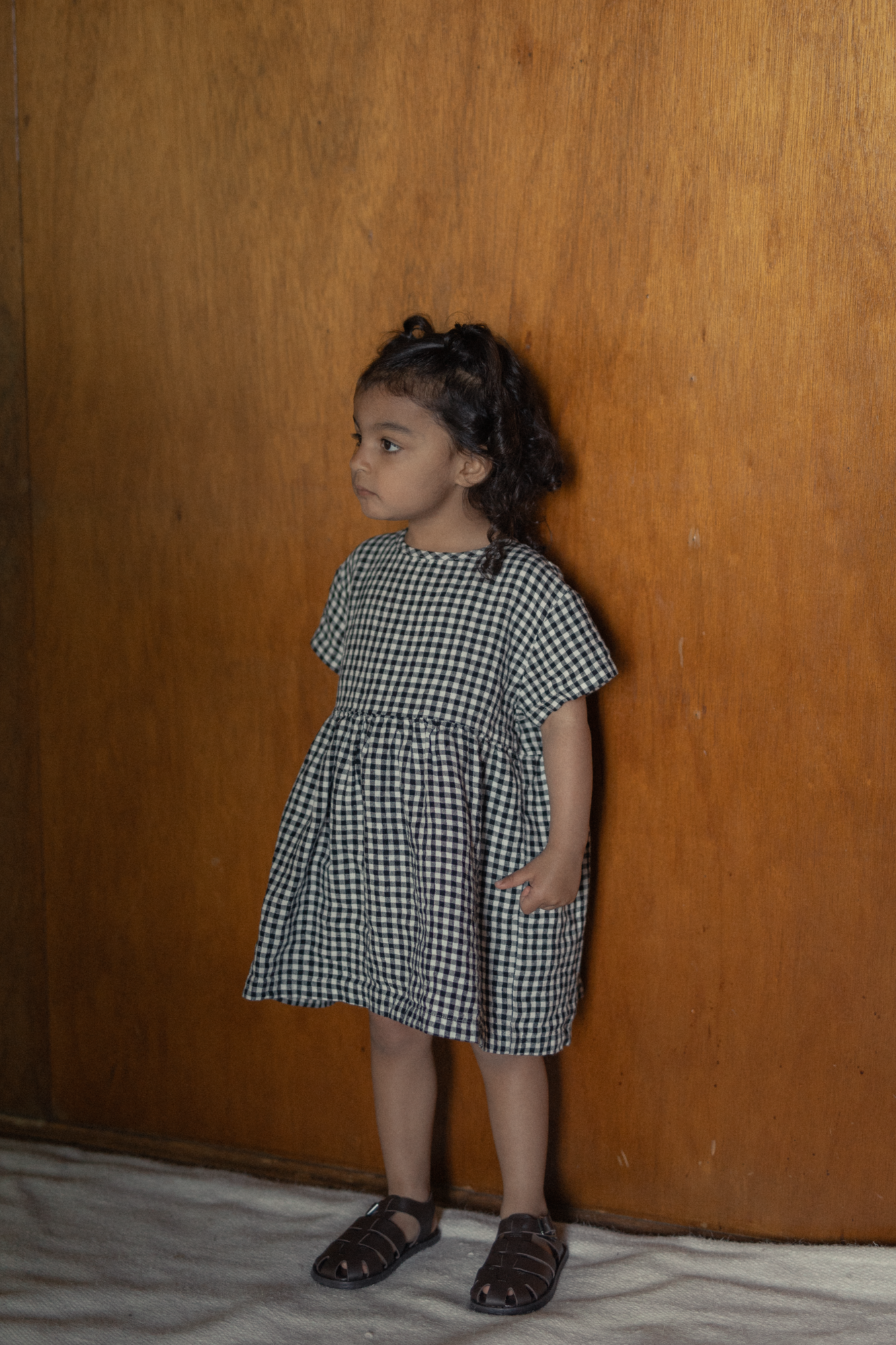 Child wearing the Mini Aya in Gingham Noir by Gillian Stevens, front view with wooden panel backdrop