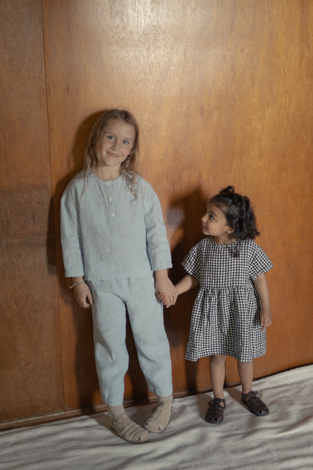 Child wearing the Mini Aya Gingham in Noir by Gillian Stevens, full-length front view with a wooden panel backdrop