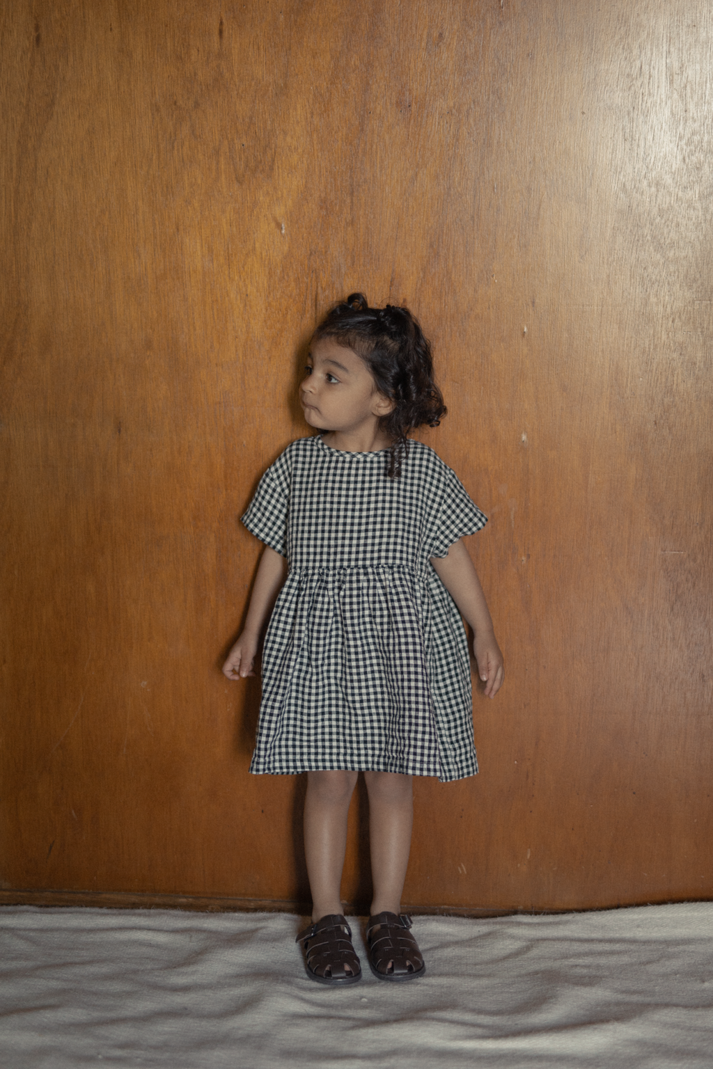 Child wearing the Mini Aya Gingham in Noir by Gillian Stevens, front view against a warm timber wall