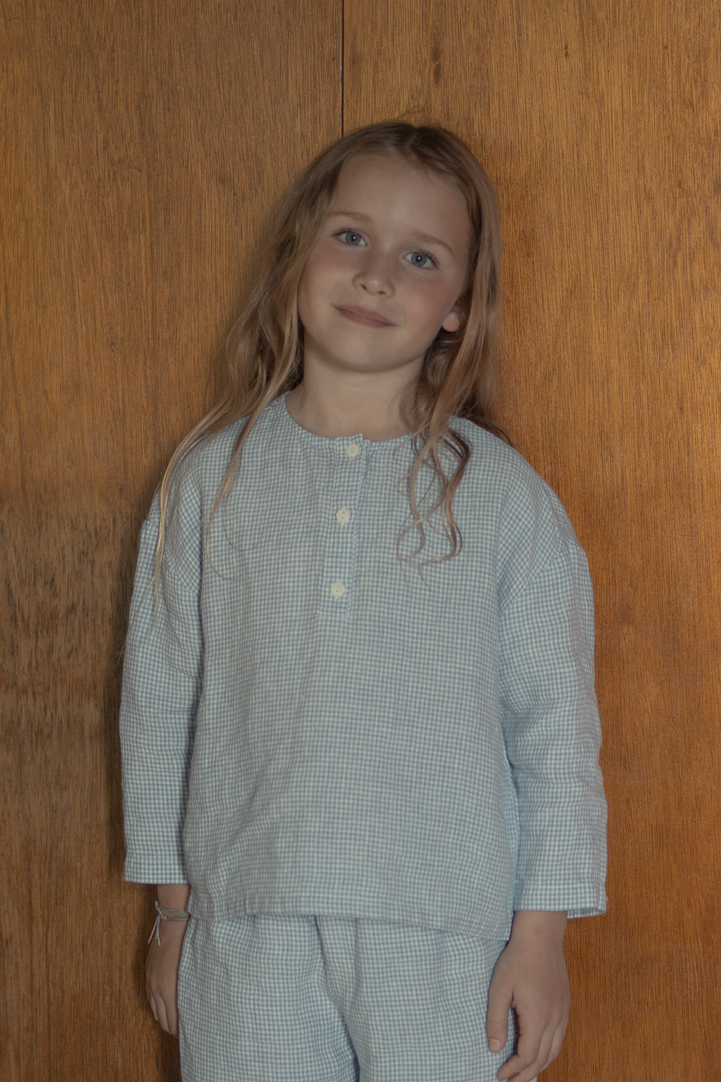 Child wearing the Bertie Top in Pale Blue Gingham by Gillian Stevens, front view against a warm timber wall