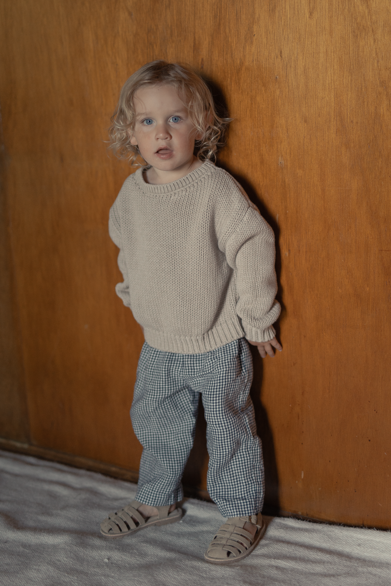 Child wearing the Mini Townes Pullover in Oat by Gillian Stevens, full-length front view against a timber wall
