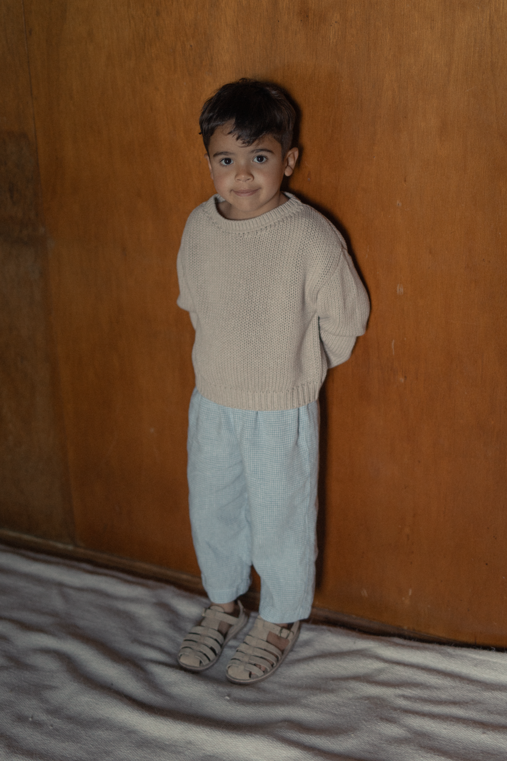 Child wearing the Mini Townes Pullover in Oat by Gillian Stevens, full-length front view against a wooden wall