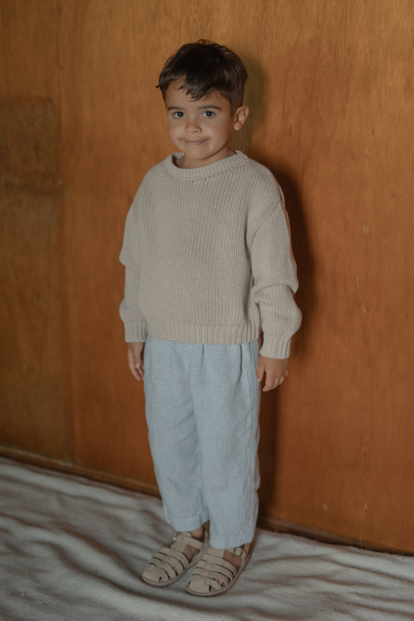 Child wearing the Mini Townes Pullover in Oat by Gillian Stevens, full-length front view against a timber wall