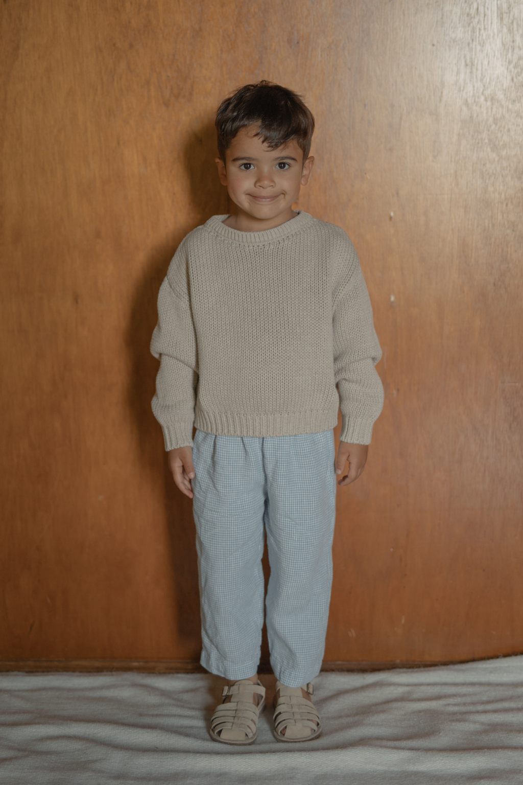 Child wearing the Bertie Pant in Pale Blue Gingham by Gillian Stevens, front view styled with a neutral knit sweater