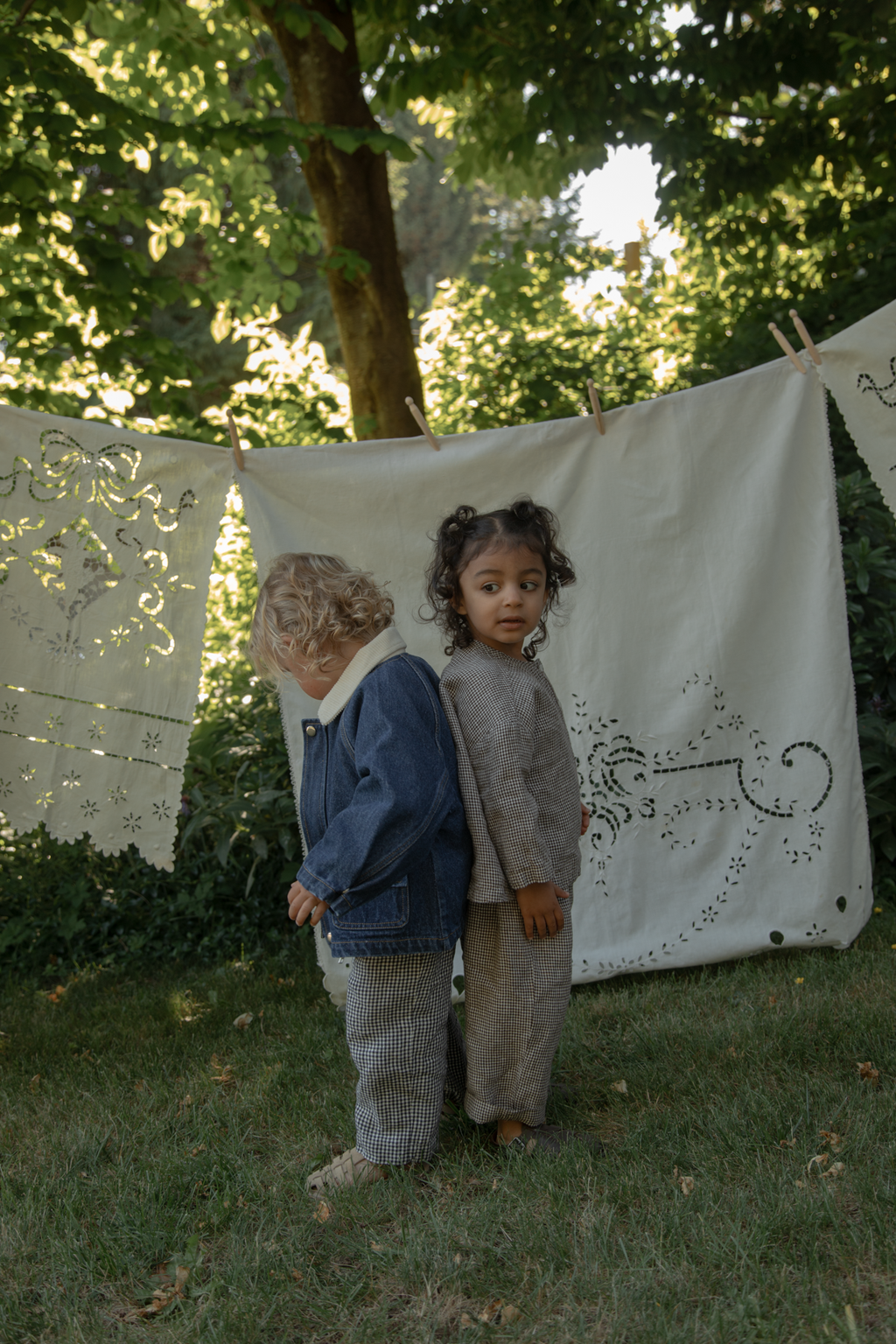 Two toddlers outdoors, one wearing the Brooks Jacket in Indigo by Gillian Stevens, back view in a garden setting