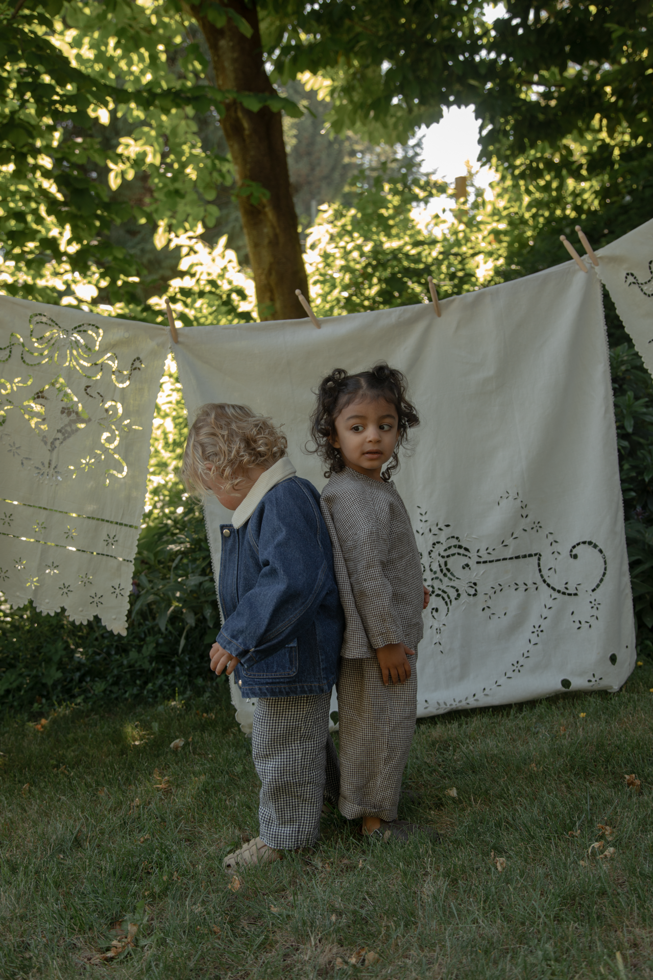 Two toddlers outdoors, one wearing the Brooks Jacket in Indigo by Gillian Stevens, back view in a garden setting