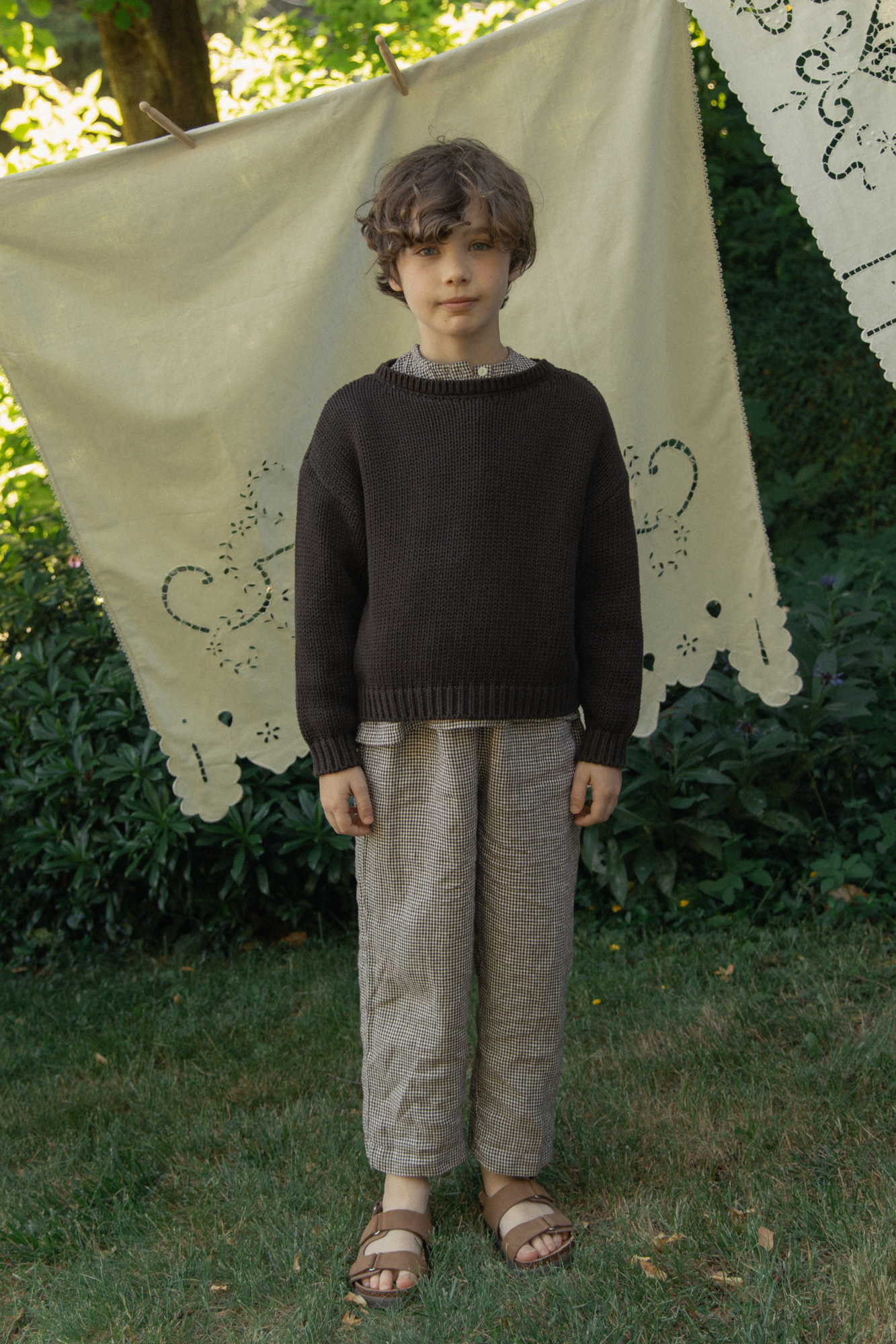 Child wearing the Mini Townes Pullover in Espresso by Gillian Stevens, front view in a garden setting