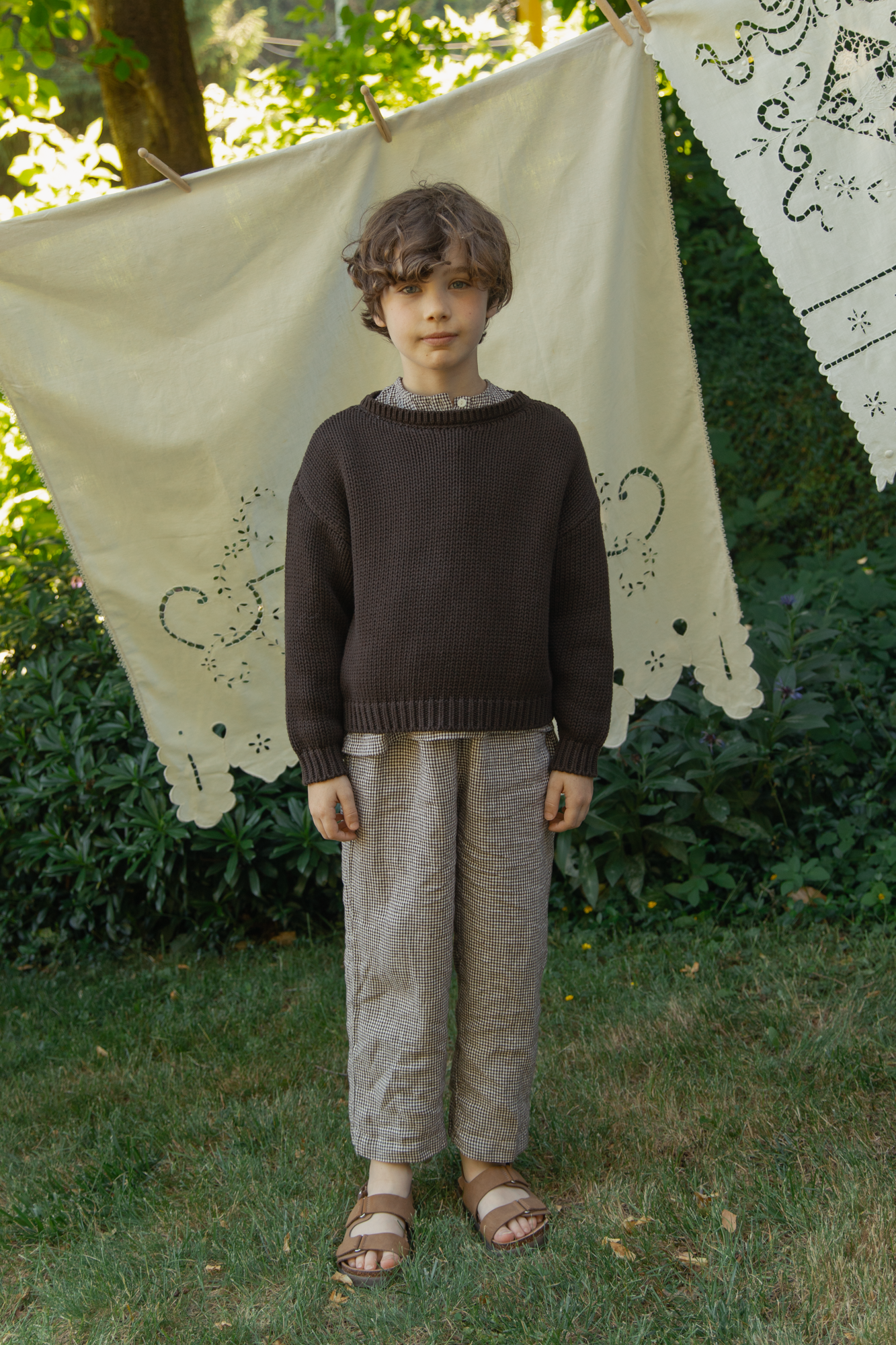 Child wearing the Mini Townes Pullover in Espresso by Gillian Stevens, front view in a garden setting