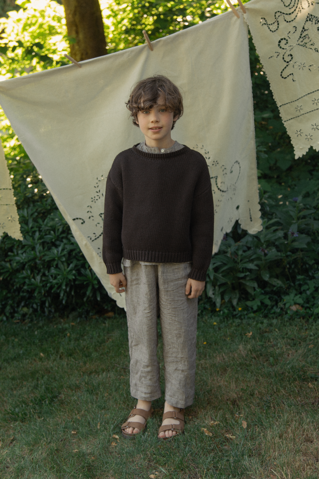 Child wearing the Mini Townes Pullover in Espresso by Gillian Stevens, front view in a garden setting