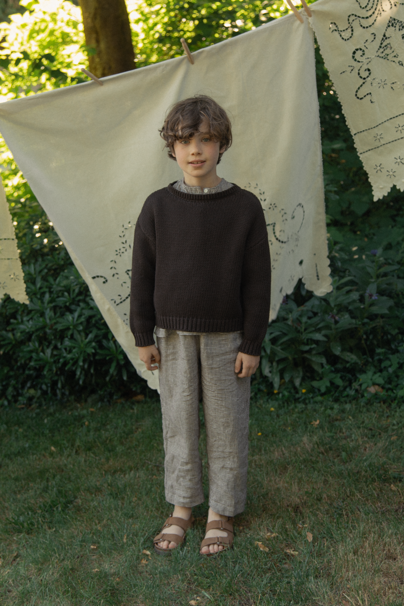 Child wearing the Mini Townes Pullover in Espresso by Gillian Stevens, front view in a garden setting