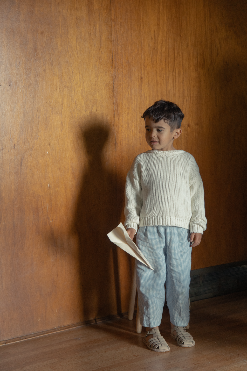 Child wearing the Mini Townes Pullover in Alabaster by Gillian Stevens, full-length front view against a wood-panelled wall