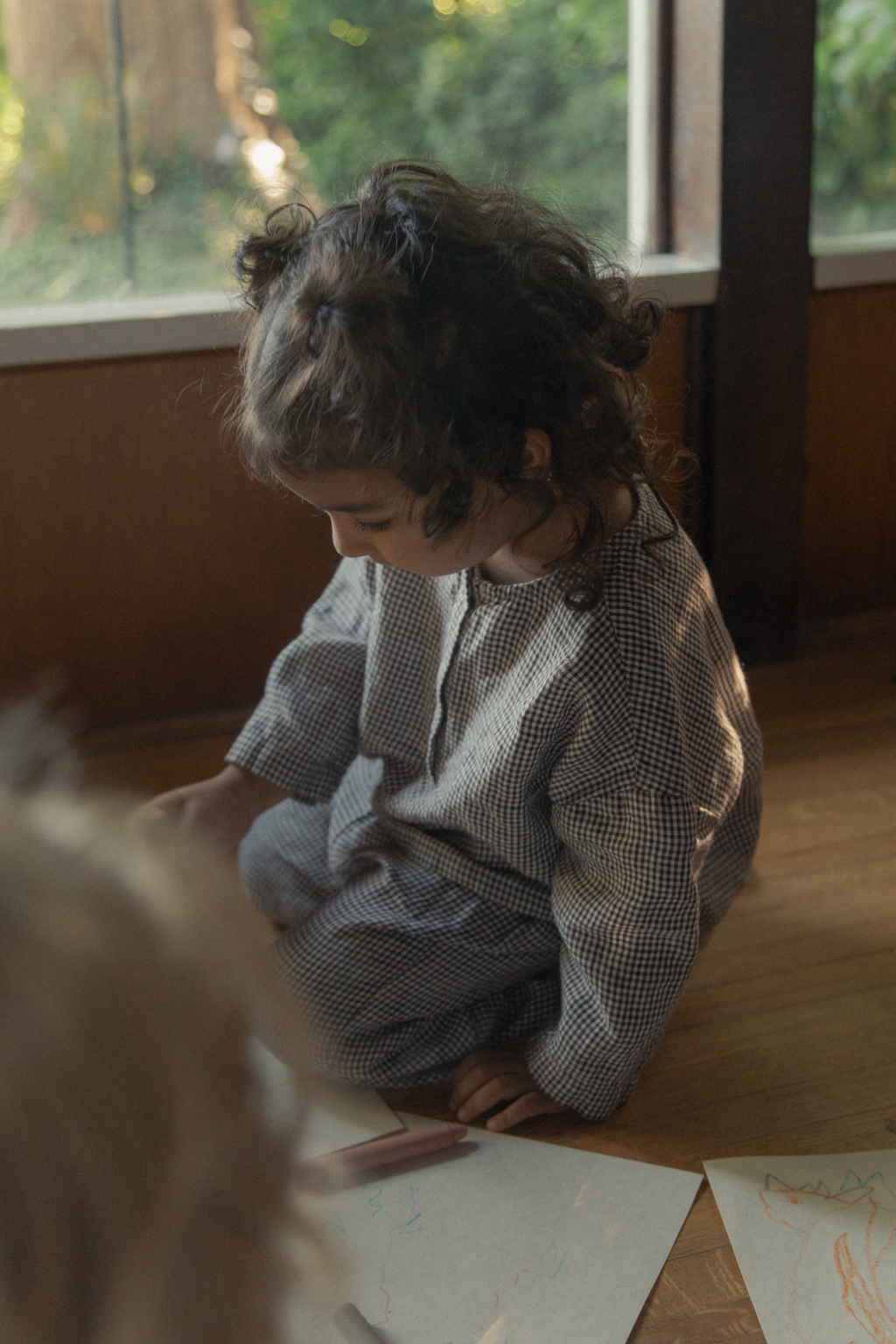 Child seated on a wooden floor in the Bertie Top Espresso Gingham by Gillian Stevens, drawing with crayons
