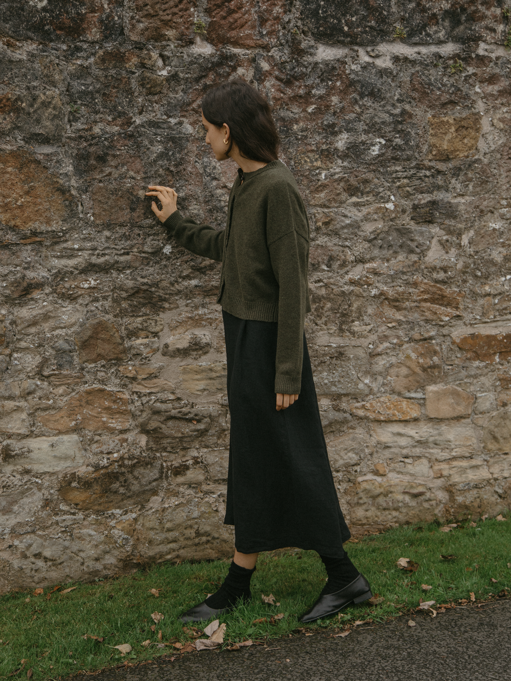 Model wearing the Giséle Cardigan in Moss by Gillian Stevens, side view against stone wall