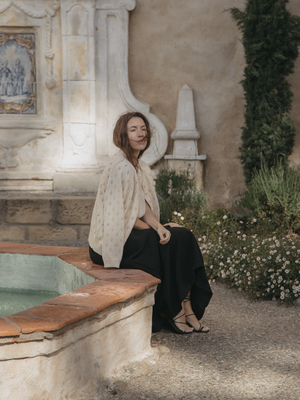 Model seated by a stone fountain wearing the Mabel Cardigan in Ivory by Gillian Stevens