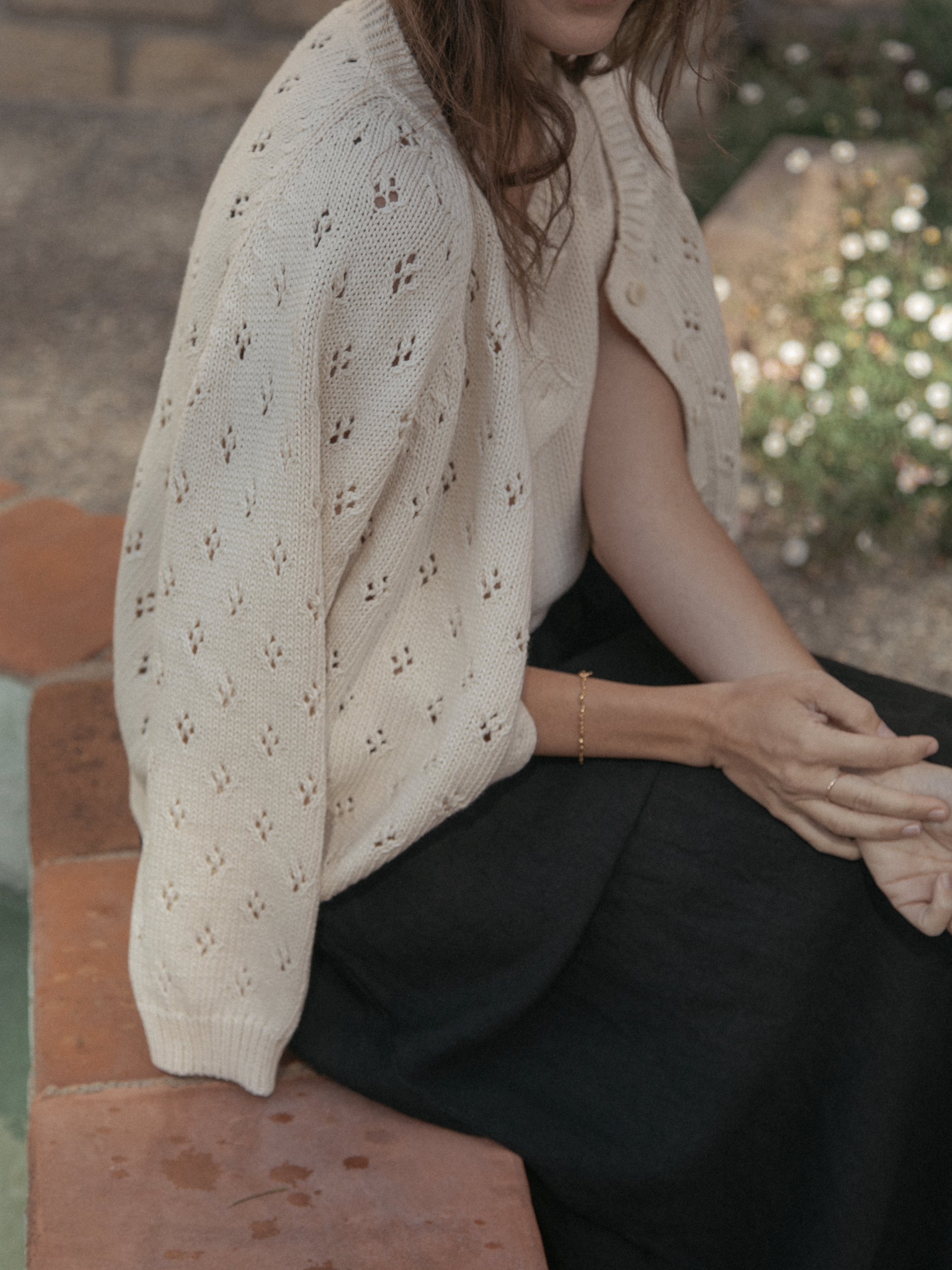 Model seated outdoors wearing the Mabel Cardigan in Ivory by Gillian Stevens, showing pointelle sleeve detail
