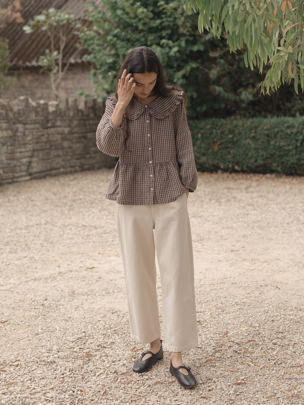 Model wearing the Winsley Blouse in Chocolate Plaid by Gillian Stevens, front view in a garden courtyard