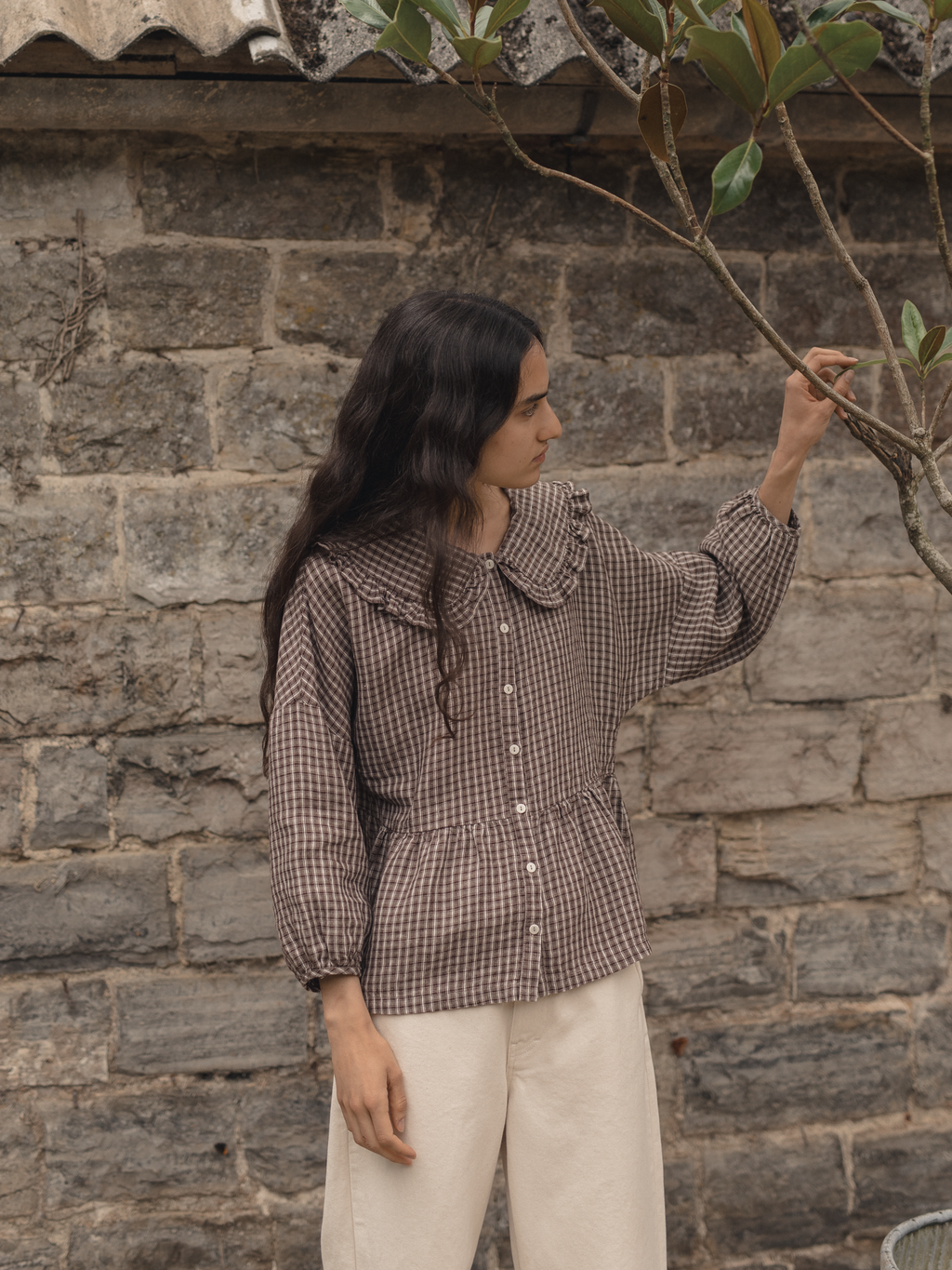 Model wearing the Winsley Blouse in Chocolate Plaid by Gillian Stevens, three-quarter front view against a stone wall