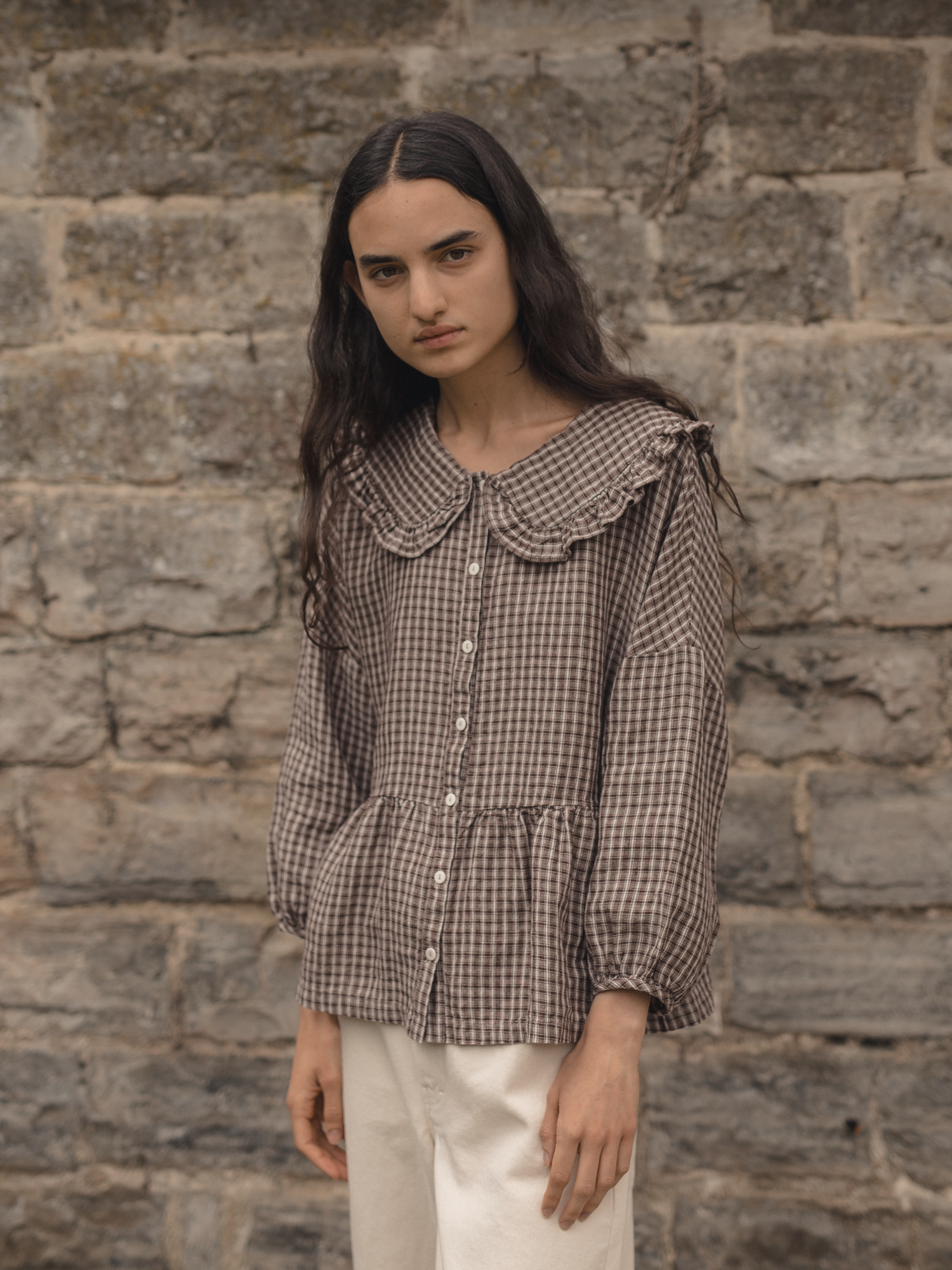 Model wearing the Winsley Blouse in Chocolate Plaid by Gillian Stevens, front view against a stone wall
