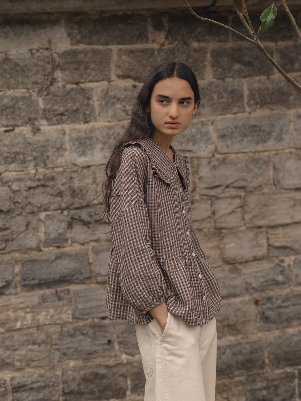 Model wearing the Winsley Blouse in Chocolate Plaid by Gillian Stevens, three-quarter view against a stone wall