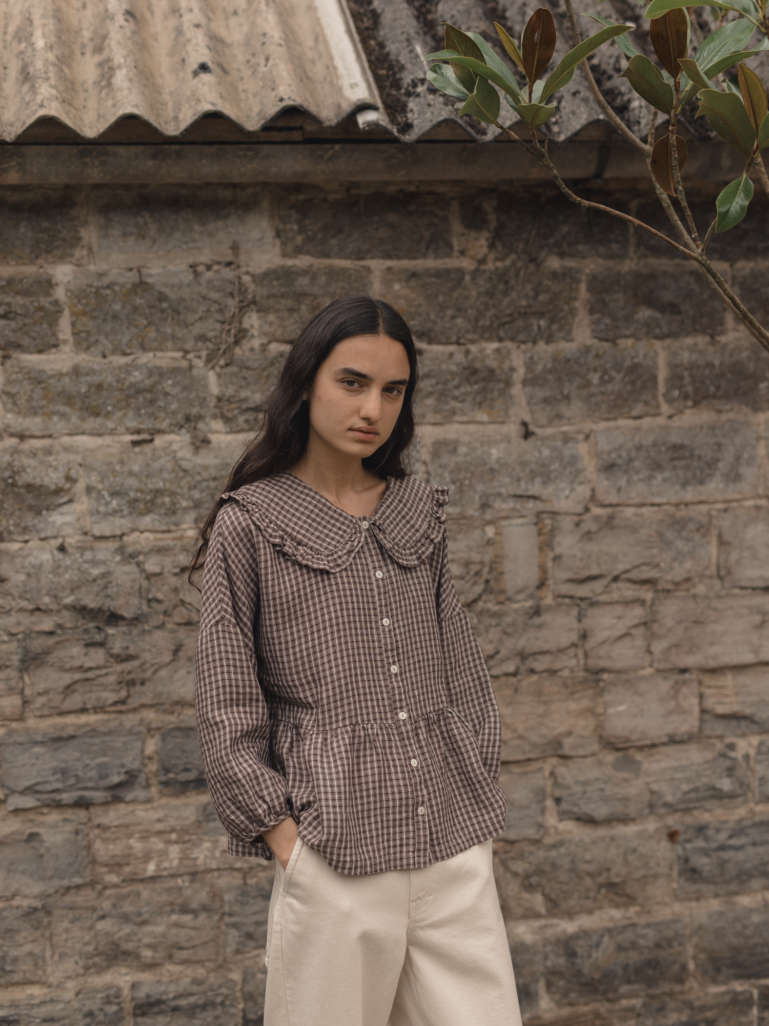 Model wearing the Winsley Blouse in Chocolate Plaid by Gillian Stevens, front view against a stone wall
