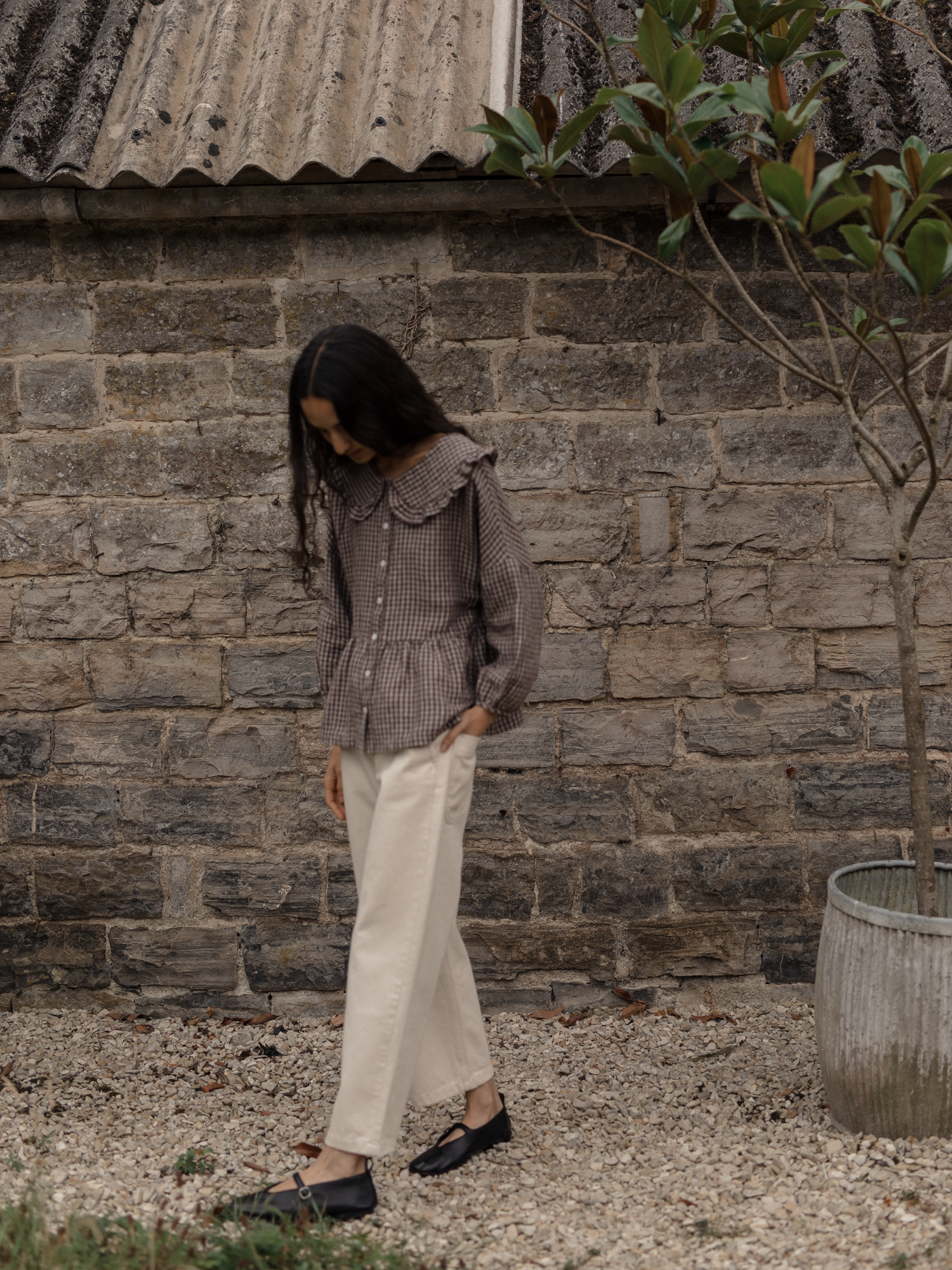 Model wearing the Winsley Blouse in Chocolate Plaid by Gillian Stevens, full-length outdoor view against a stone wall