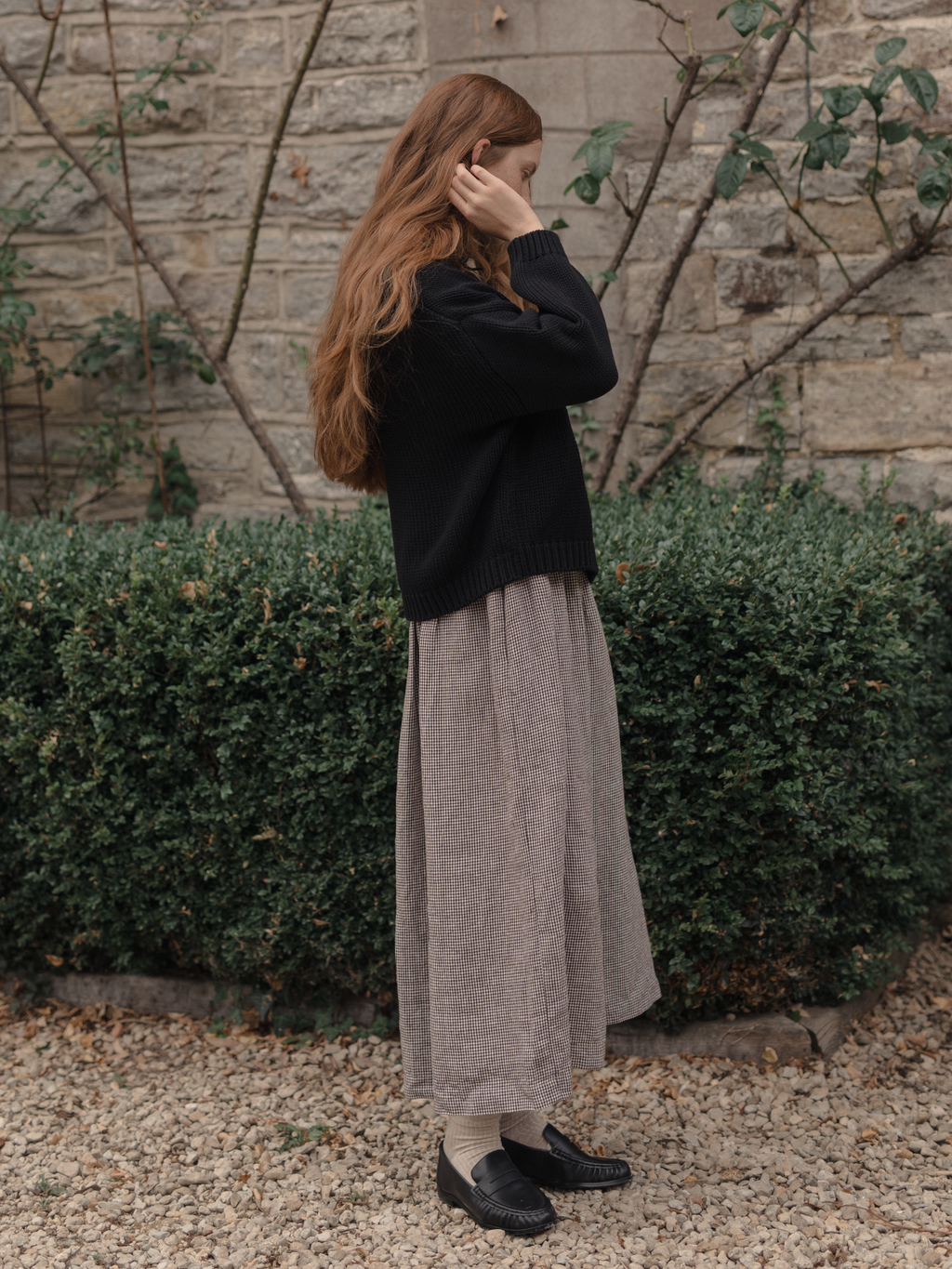 Model wearing the Townes Pullover in Noir by Gillian Stevens, side view styled with a check midi skirt