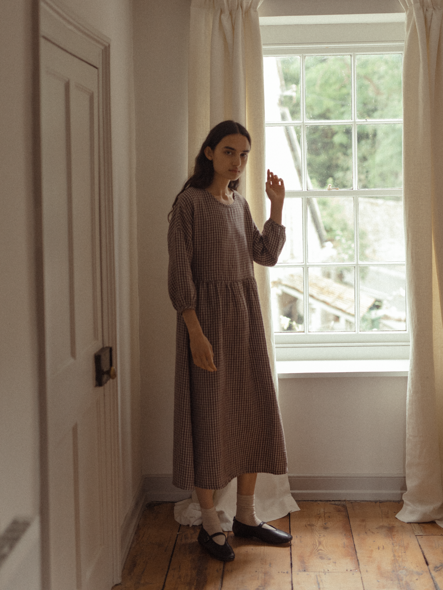 Model wearing the Darla Dress in Chocolate Plaid by Gillian Stevens, front view beside a sunlit window
