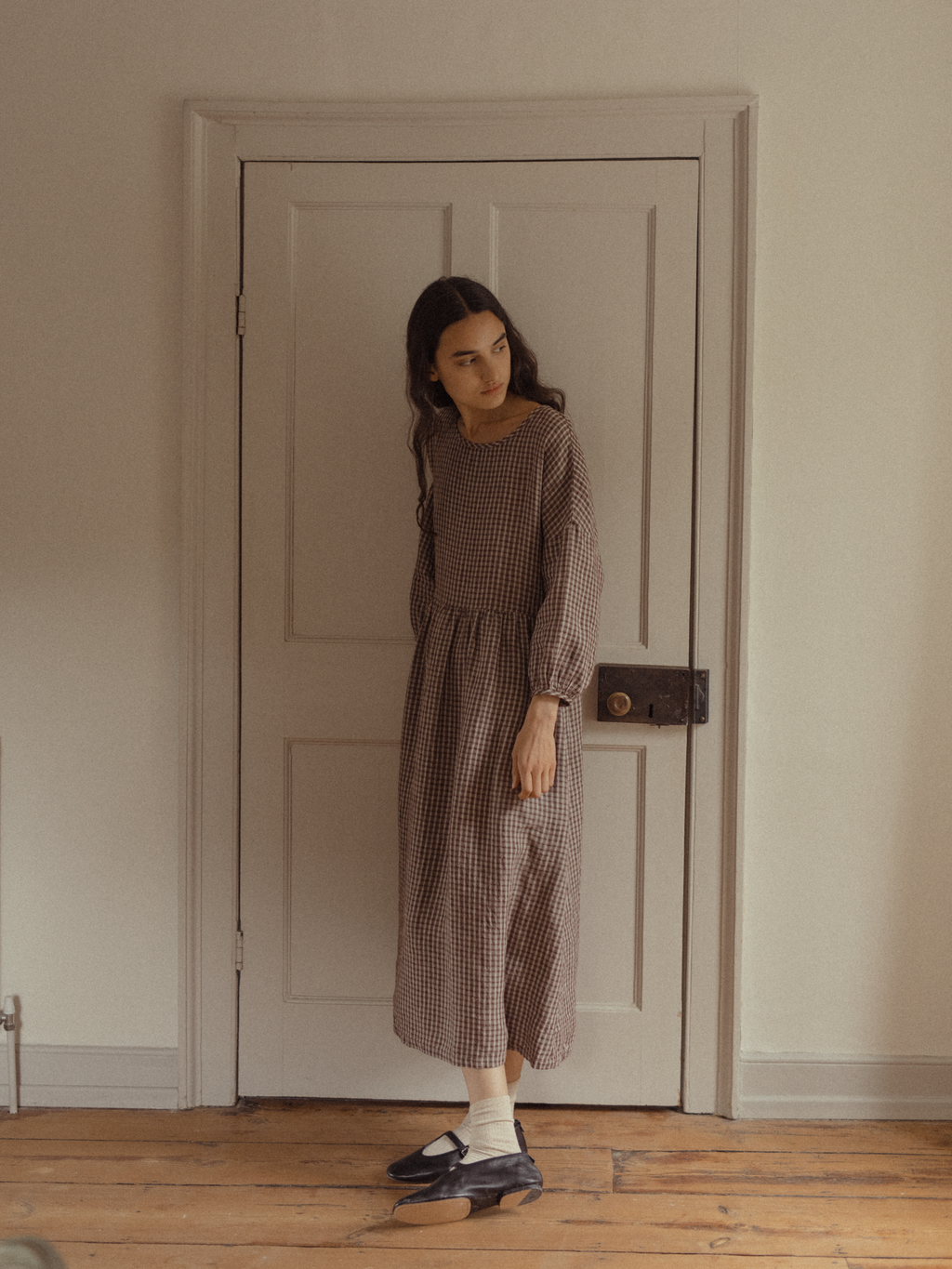 Model wearing the Darla Dress in Chocolate Plaid by Gillian Stevens, full-length front view against a vintage door