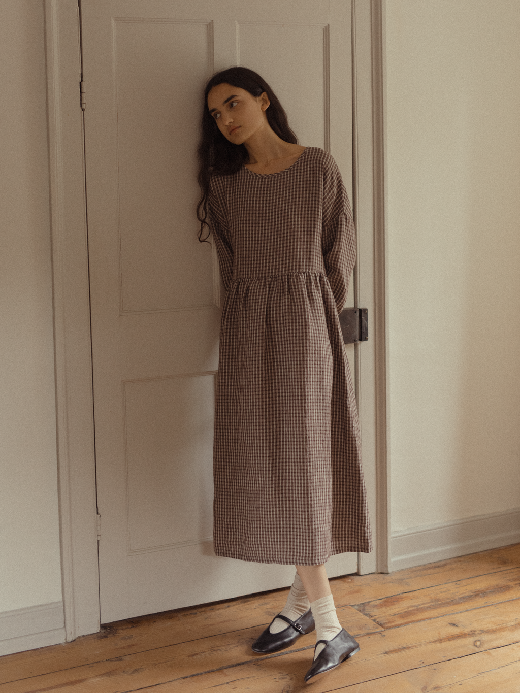 Model wearing the Darla Dress in Chocolate Plaid by Gillian Stevens, front view leaning against a door