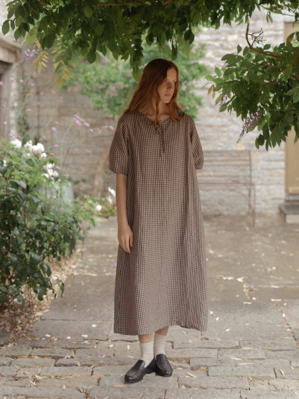 Model wearing the Blair Dress in Chocolate Plaid by Gillian Stevens, front view in a garden courtyard