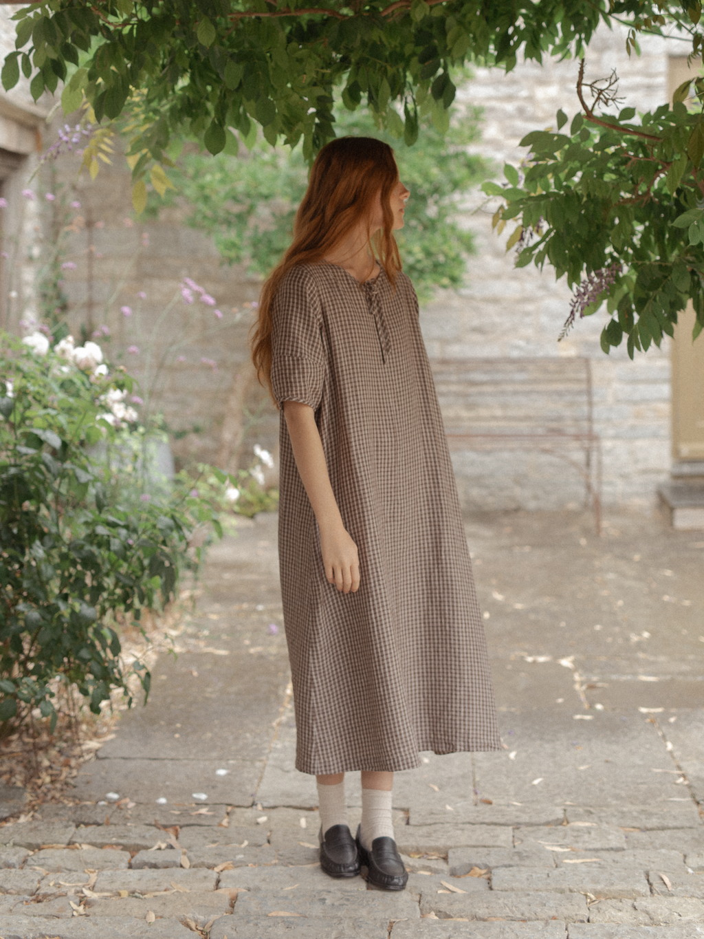 Model wearing the Blair Dress in Chocolate Plaid by Gillian Stevens, three-quarter side view in a stone courtyard garden