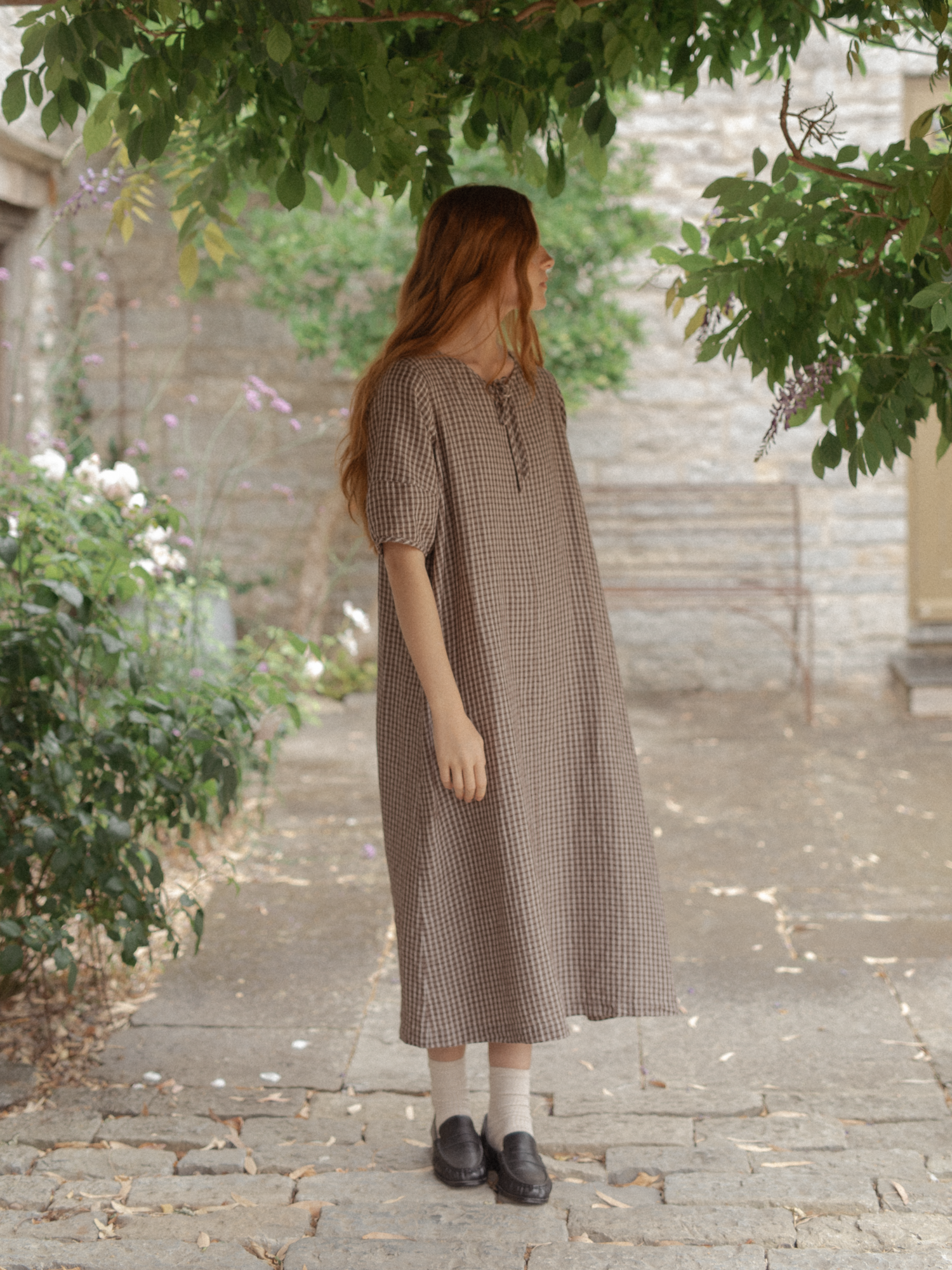 Model wearing the Blair Dress in Chocolate Plaid by Gillian Stevens, three-quarter side view in a stone courtyard garden