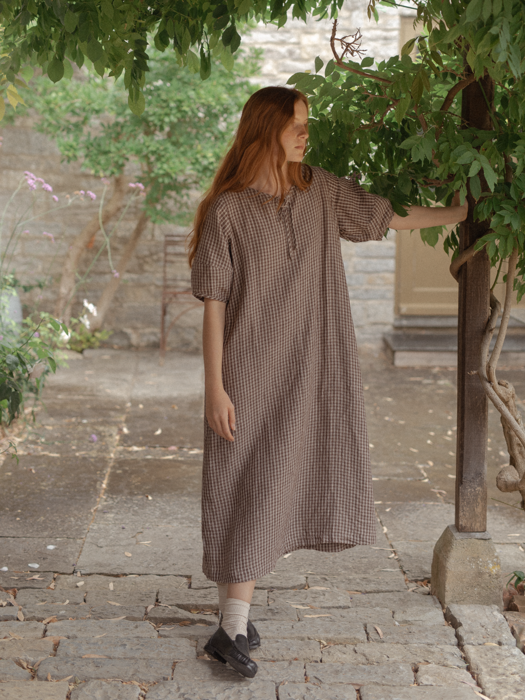 Model wearing the Blair Dress in Chocolate Plaid by Gillian Stevens, front view in a stone courtyard garden
