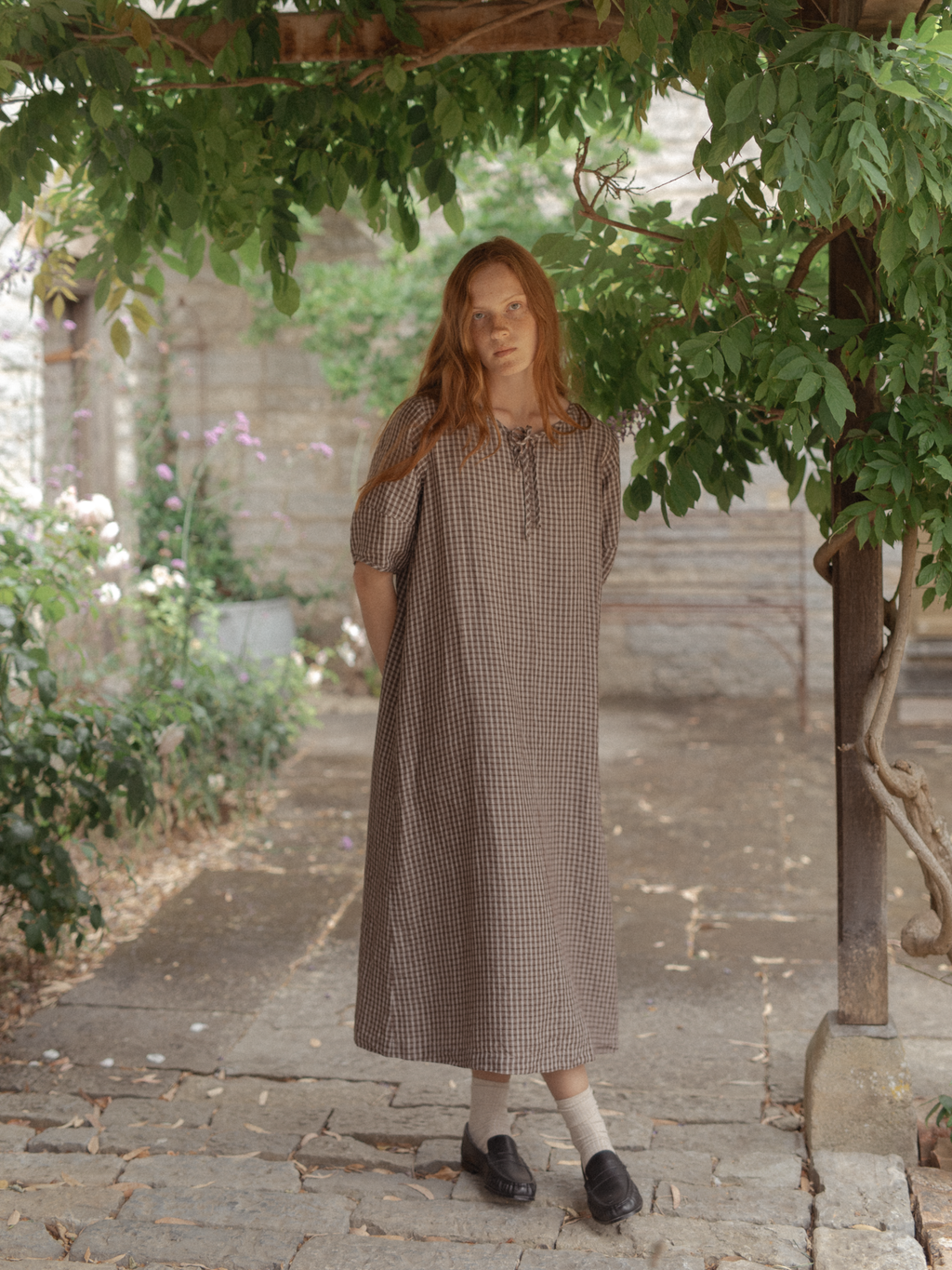 Model wearing the Blair Dress in Chocolate Plaid by Gillian Stevens, front view in a sunlit garden courtyard