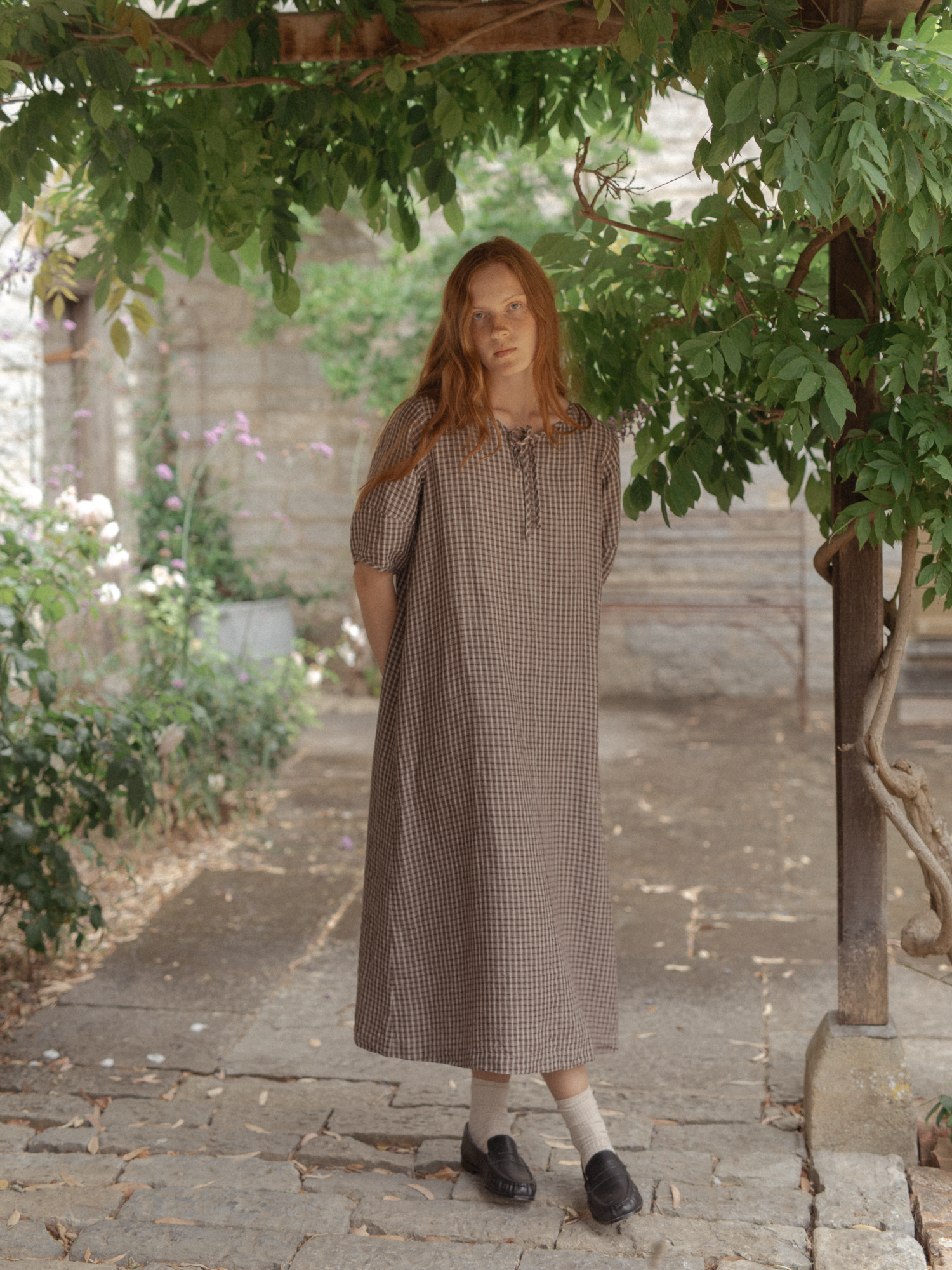 Model wearing the Blair Dress in Chocolate Plaid by Gillian Stevens, front view in a sunlit garden courtyard