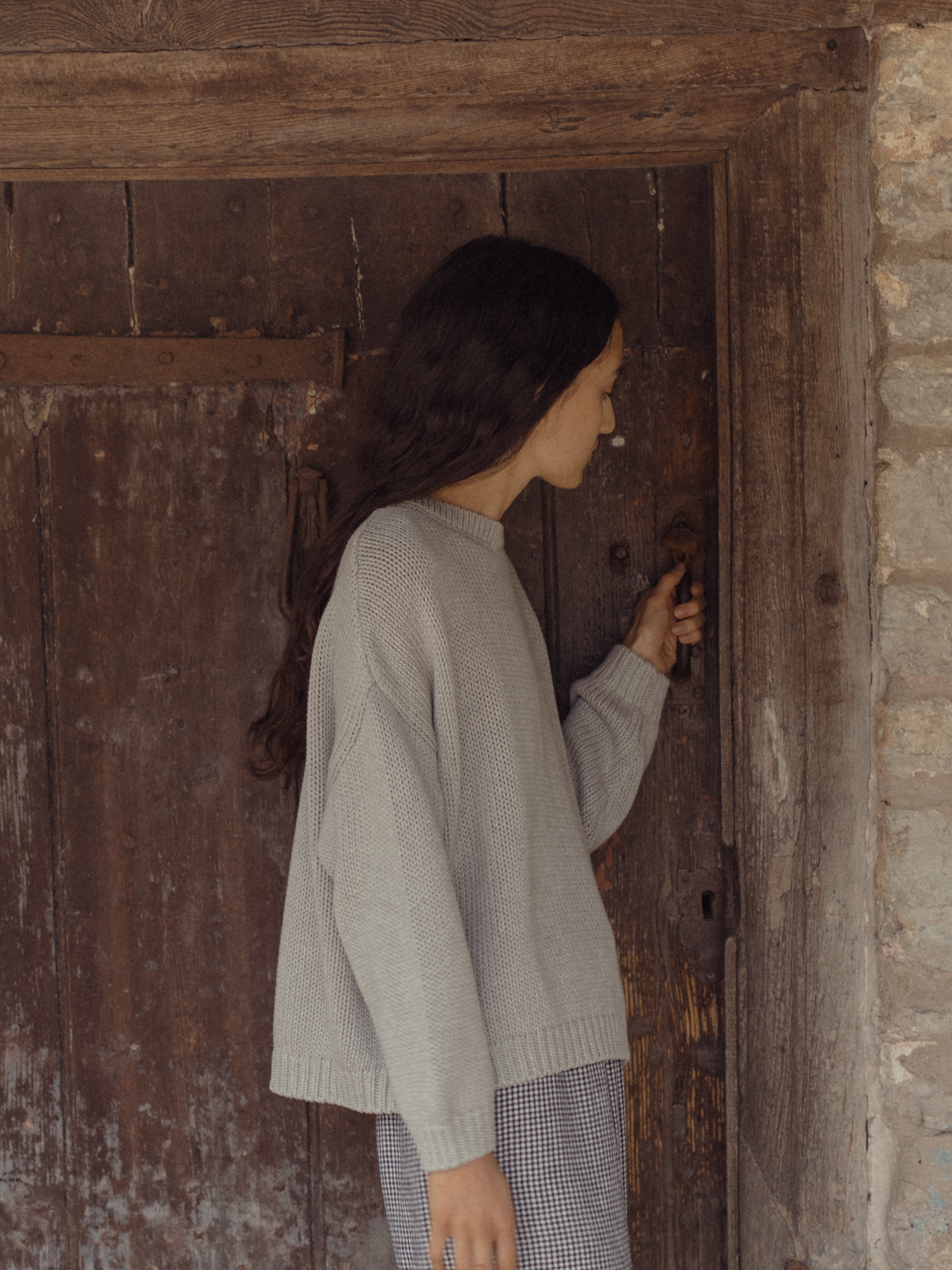 Model wearing the Townes Pullover in Pebble by Gillian Stevens, side view at a rustic wooden door