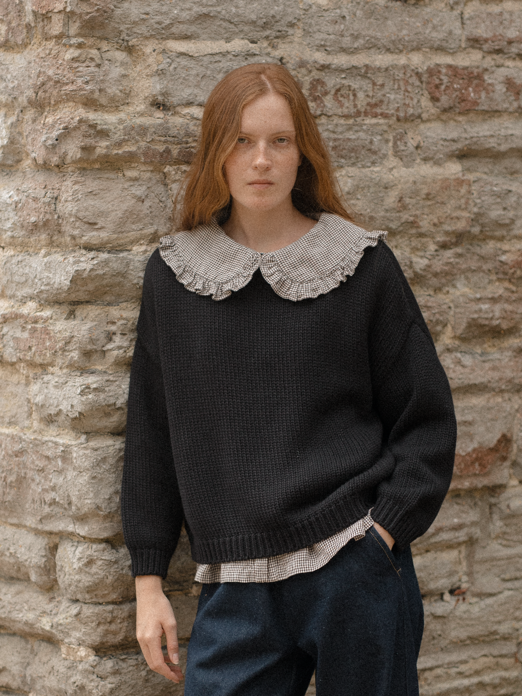 Model wearing the Townes Pullover in Noir by Gillian Stevens, styled with a gingham frill collar shirt against a stone wall