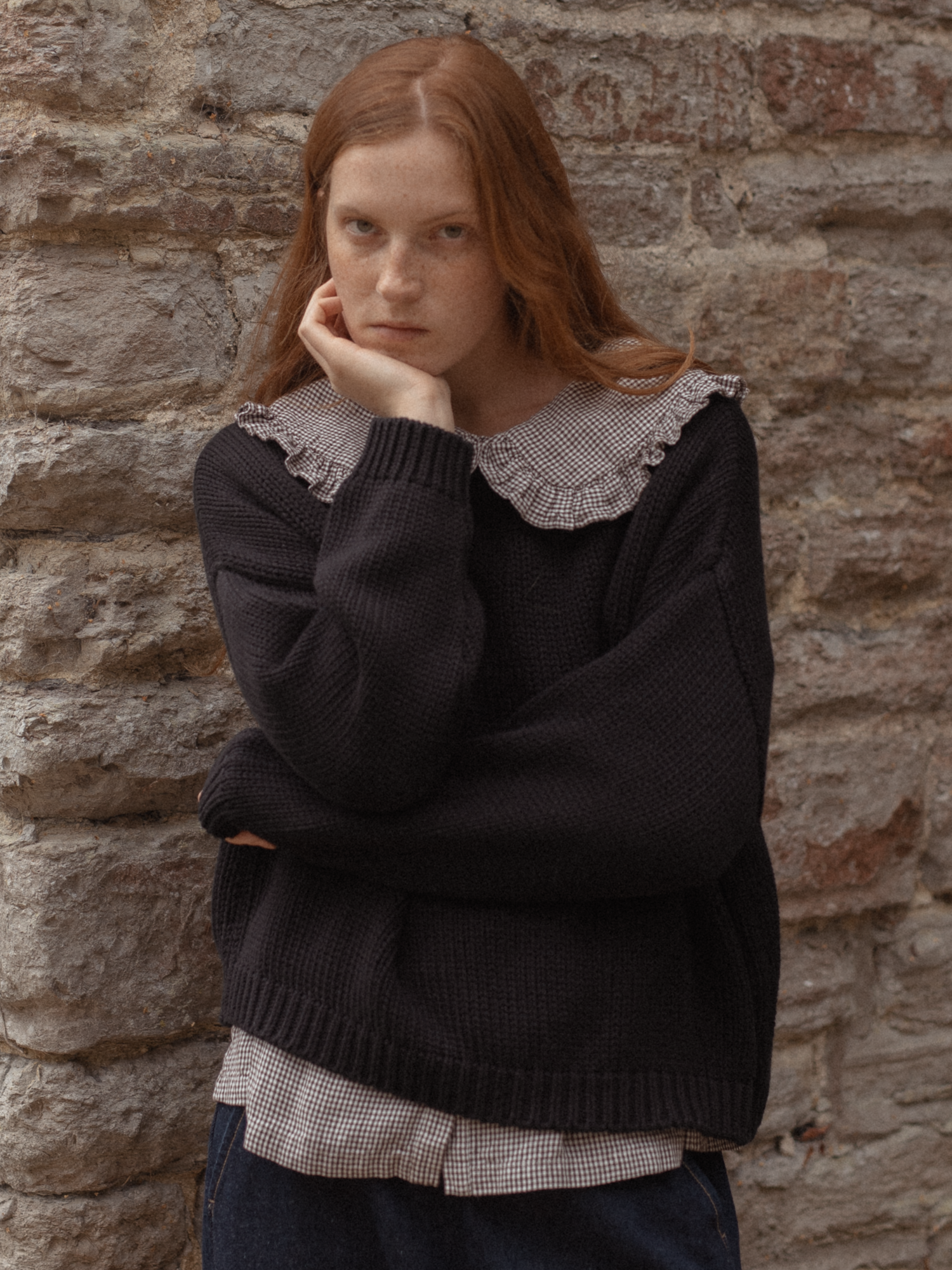 Model wearing the Townes Pullover in Noir by Gillian Stevens, layered over a gingham shirt against a stone wall