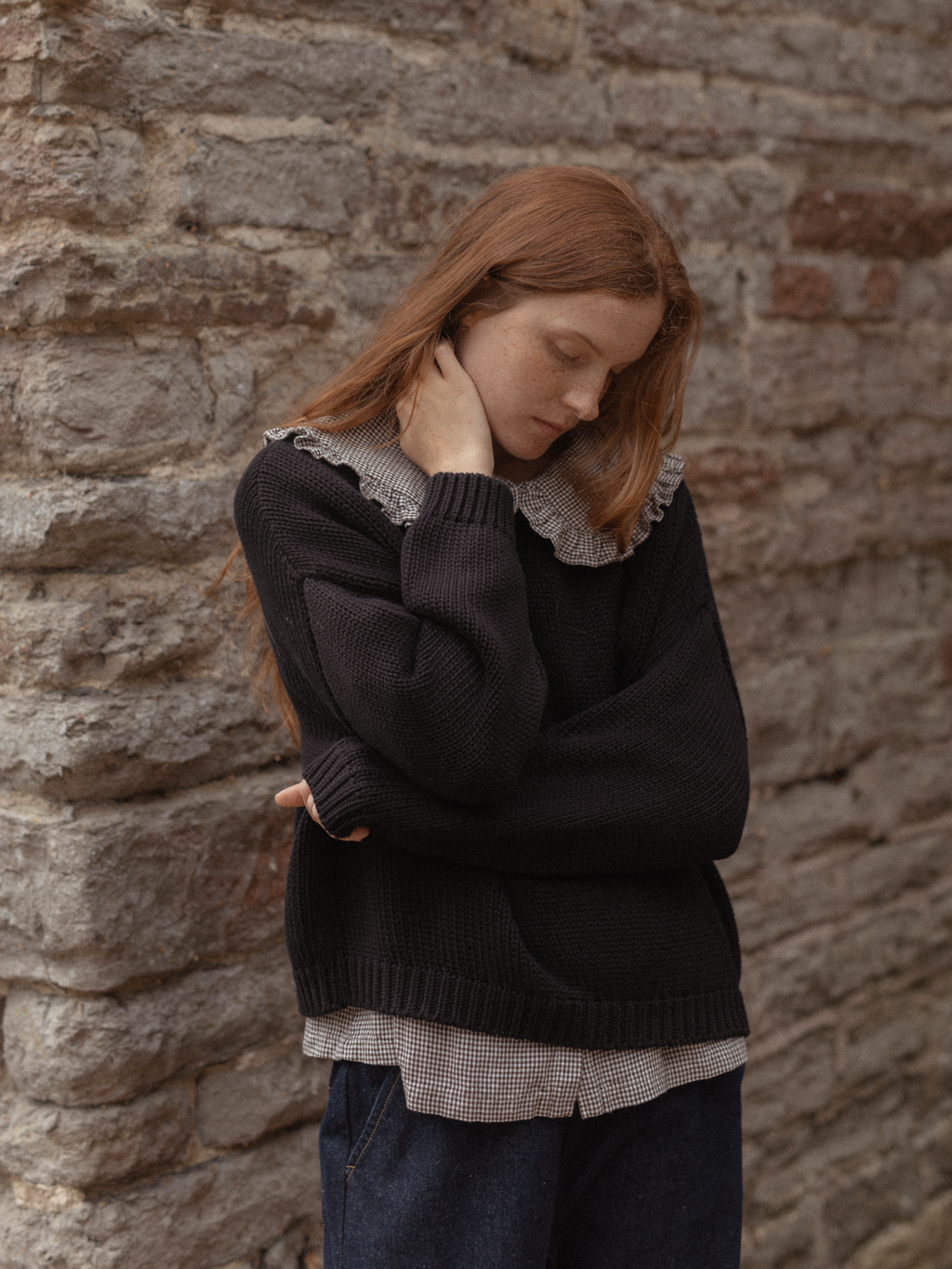 Model wearing the Townes Pullover in Noir by Gillian Stevens, layered over a gingham shirt against a stone wall