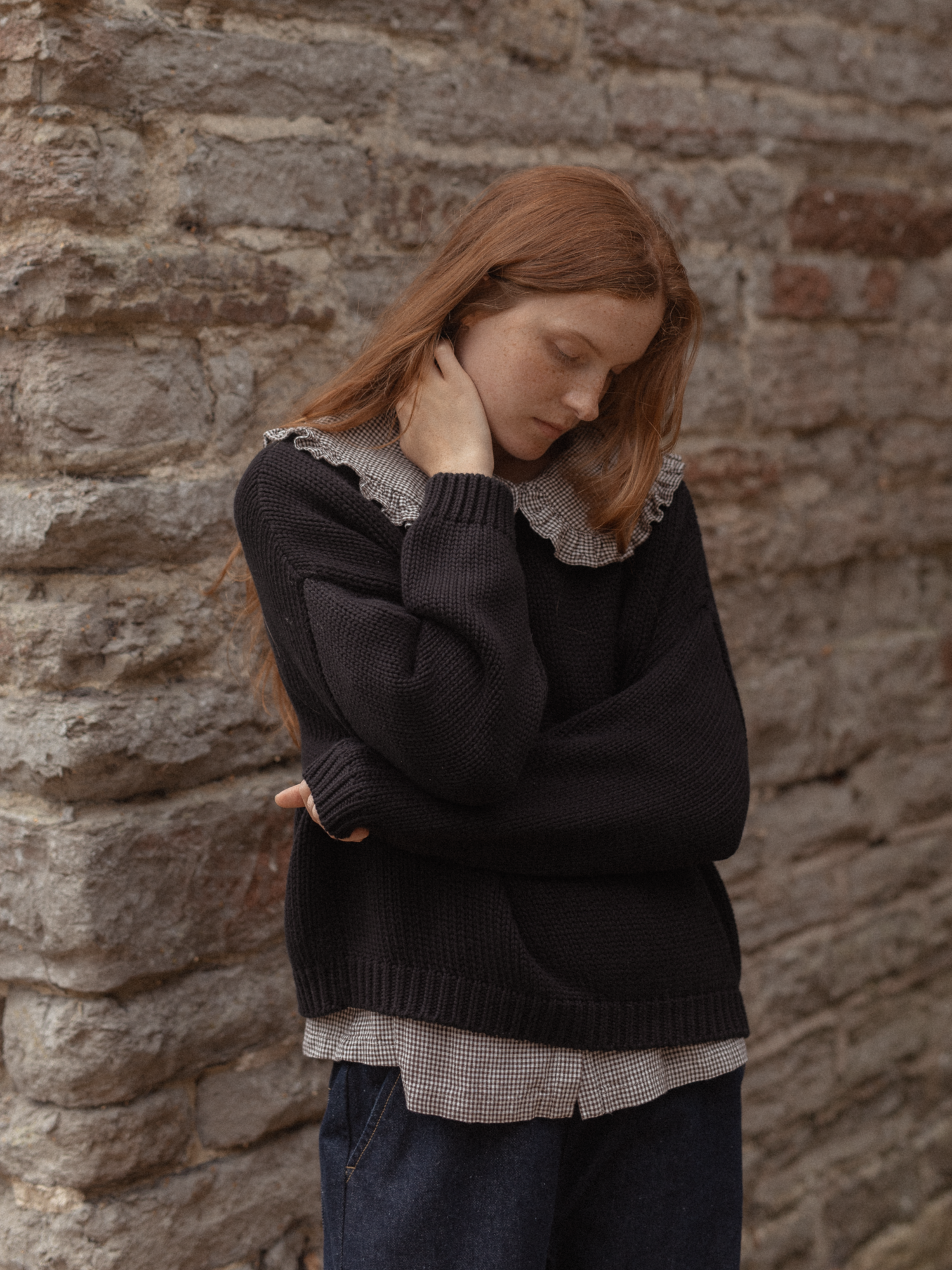 Model wearing the Townes Pullover in Noir by Gillian Stevens, layered over a gingham shirt against a stone wall