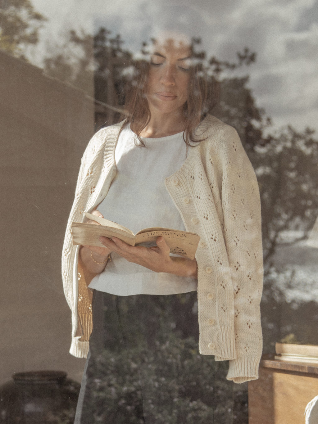 Model wearing the Mabel Cardigan in Ivory by Gillian Stevens, reading a book shot through glass