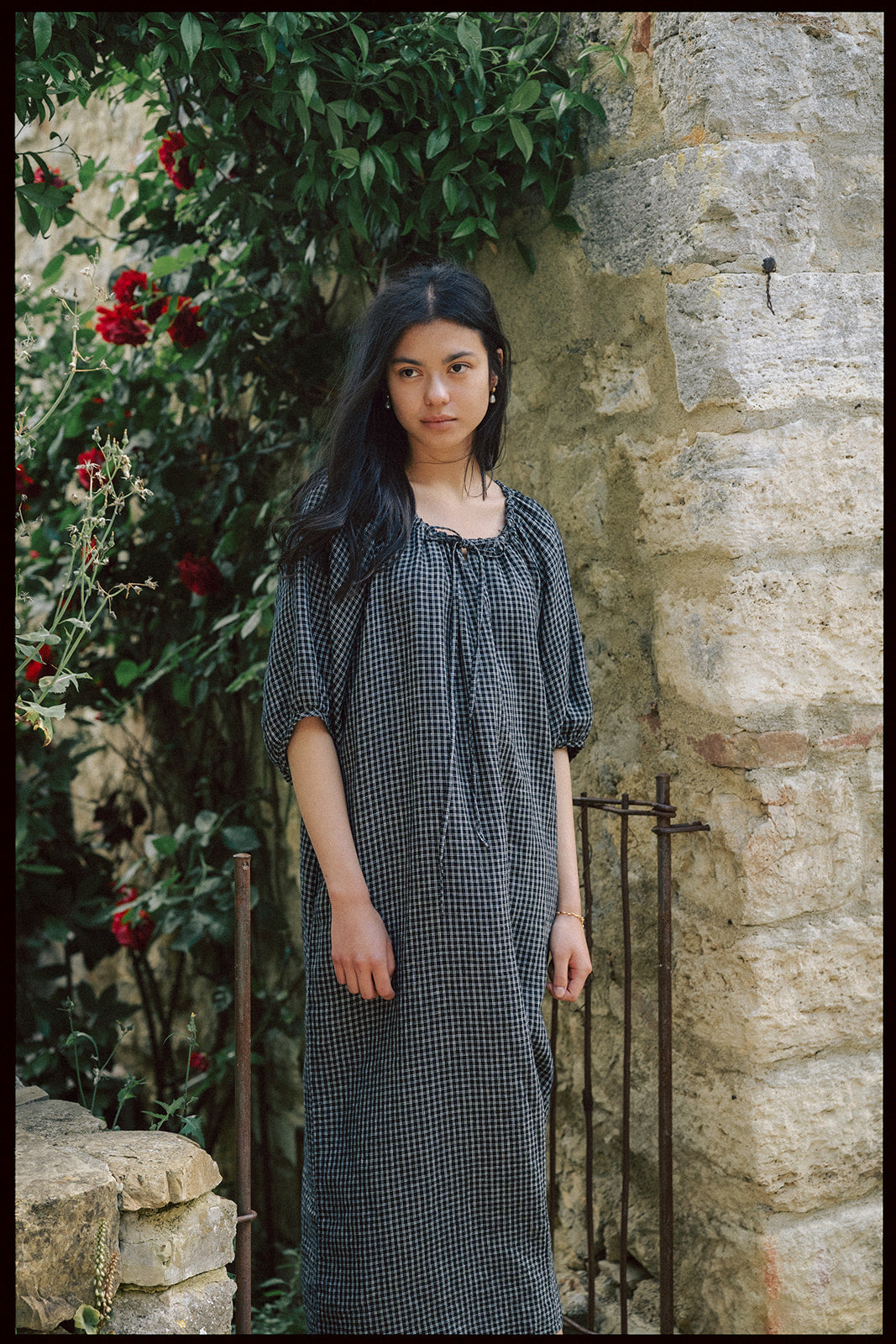 Model wearing the Jones Dress in Black Check by Gillian Stevens, front view beside a stone wall with red roses