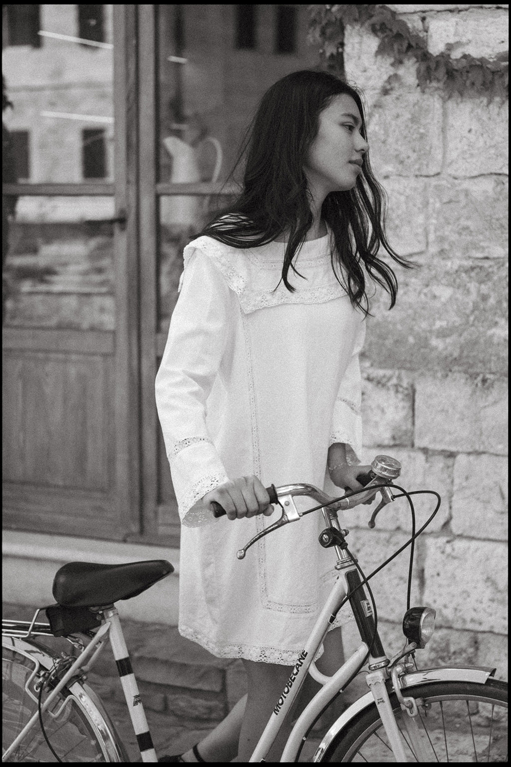 Model wearing the Laine Dress in Salt by Gillian Stevens, side profile holding a bicycle beside a stone wall