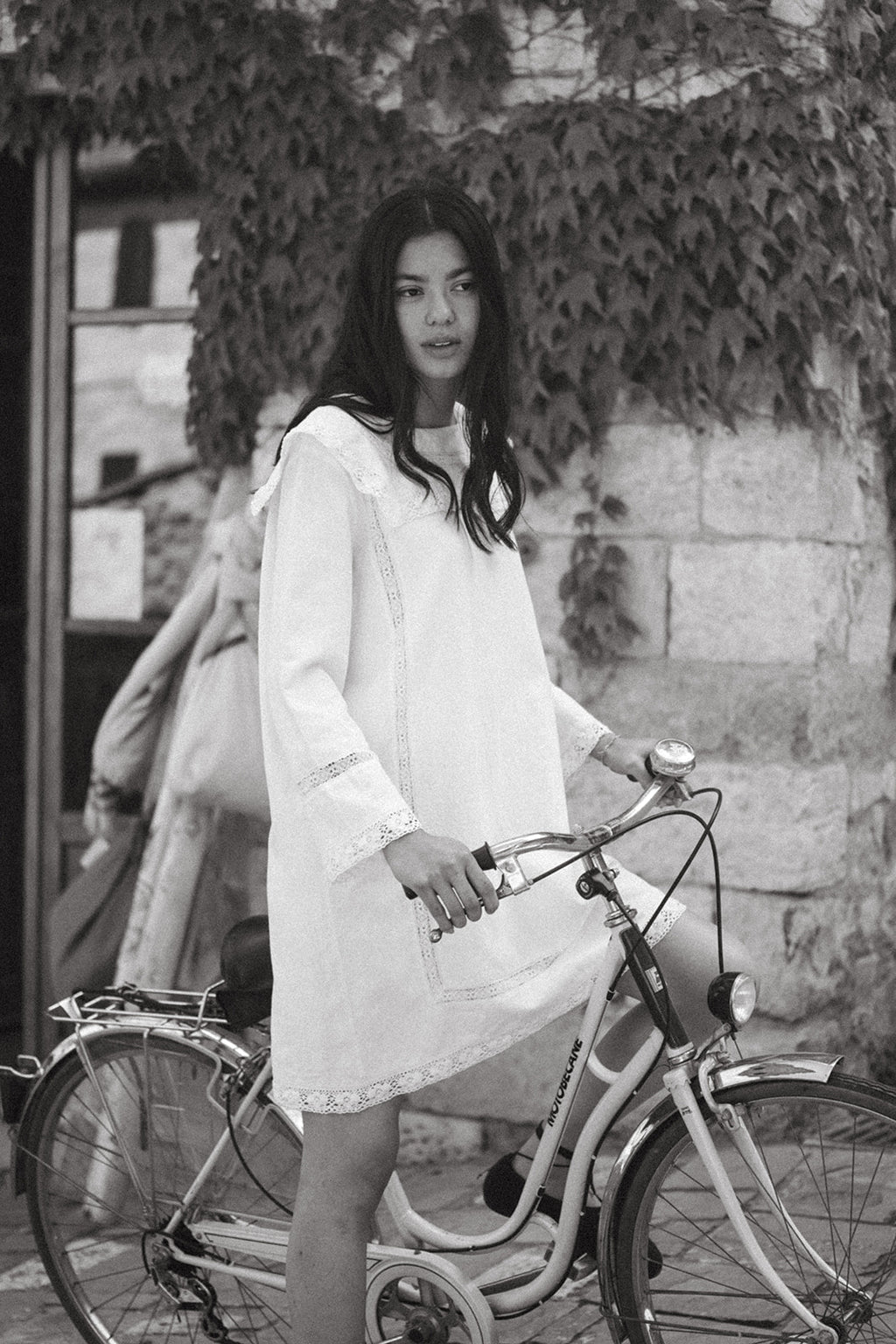 Model wearing the Laine Dress in Salt by Gillian Stevens, front view beside a vintage bicycle outdoors