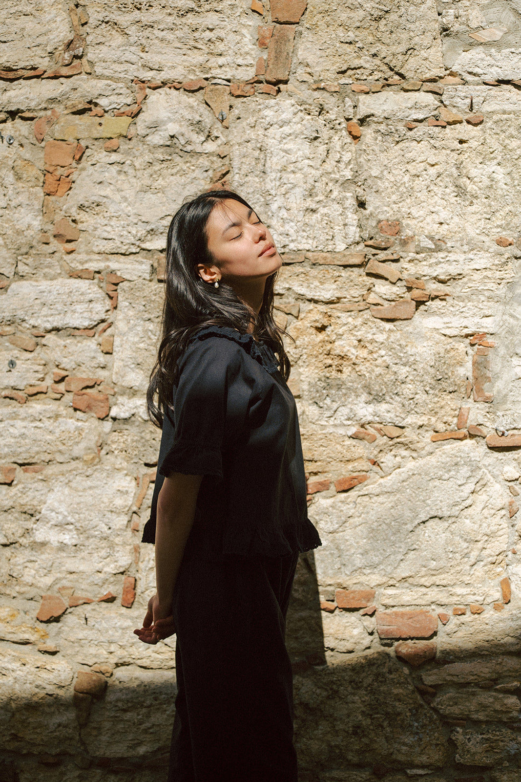 Model wearing the Arlette Top in Noir by Gillian Stevens, side profile against a rustic stone wall