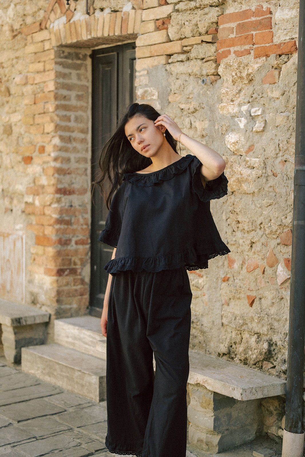 Model wearing the Arlette Top in Noir by Gillian Stevens, front view against a rustic stone wall