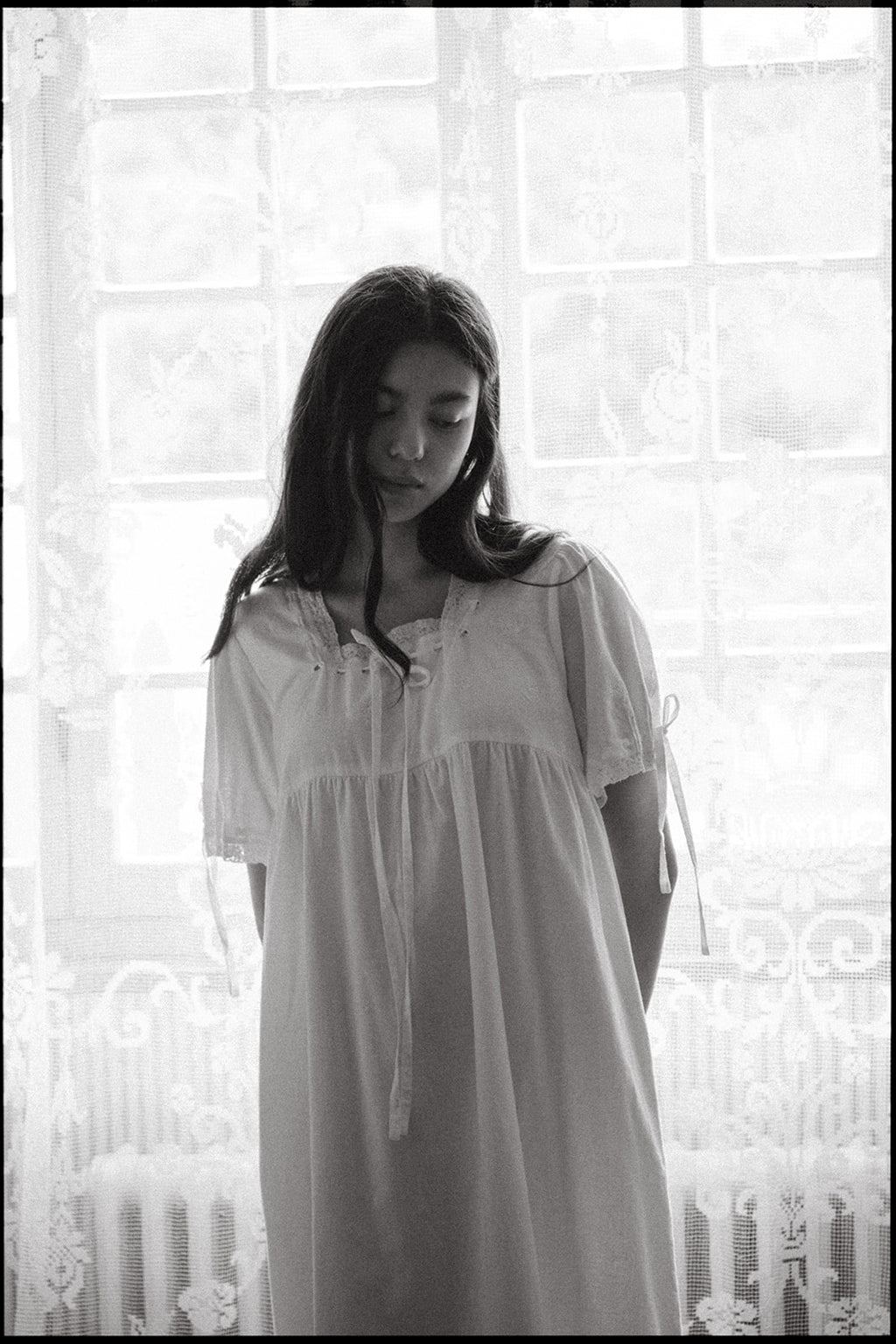 Model wearing the Dace Dress in Salt by Gillian Stevens, front view by a lace-curtained window