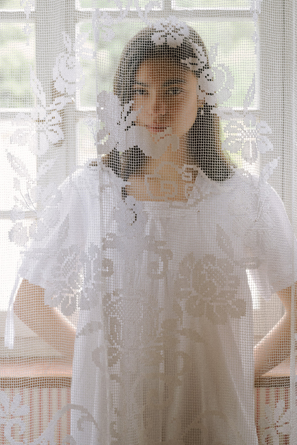 Model wearing the Dace Dress in Salt by Gillian Stevens, seen through a sheer lace curtain