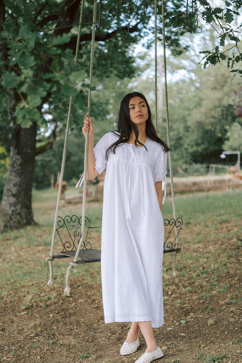 Model wearing the Dace Dress in Salt by Gillian Stevens, standing beside a rope swing in a garden setting