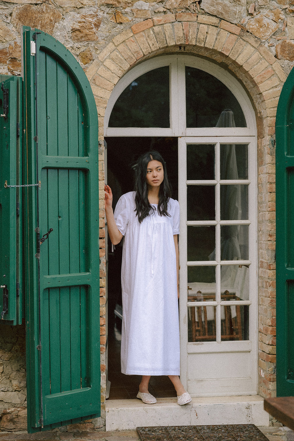 Model wearing the Dace Dress in Salt by Gillian Stevens, standing in a stone archway with green shutters